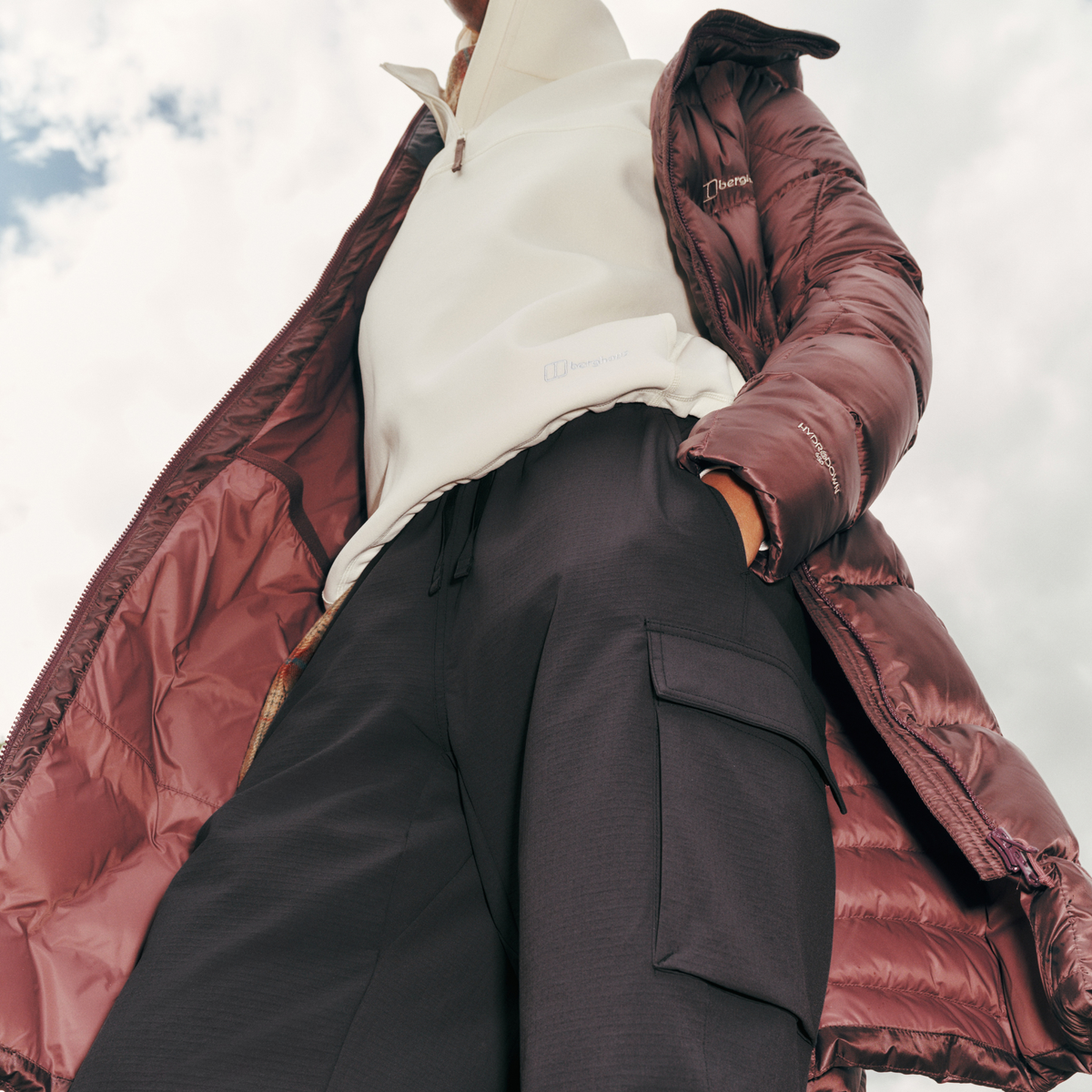 Close up view from below of a woman wearing Berghaus Women's Explorer Long Hooded Down Insulated Jacket Chakra Deep Purple 002 A002093 open, hands in her pockets