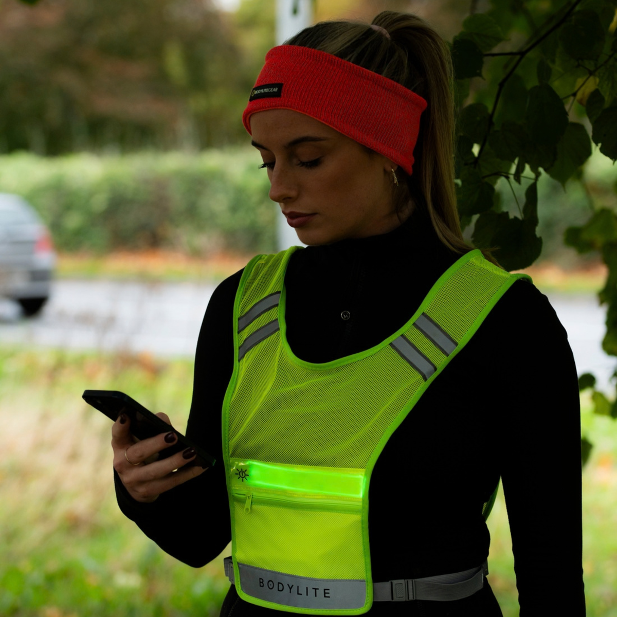 A woman outdoors wearing a Bodylite Gear Unisex V2 Reflective Vest Neon Yellow 01309-ALL SIZeS over a black top, with a pink headband and a phone in hand.