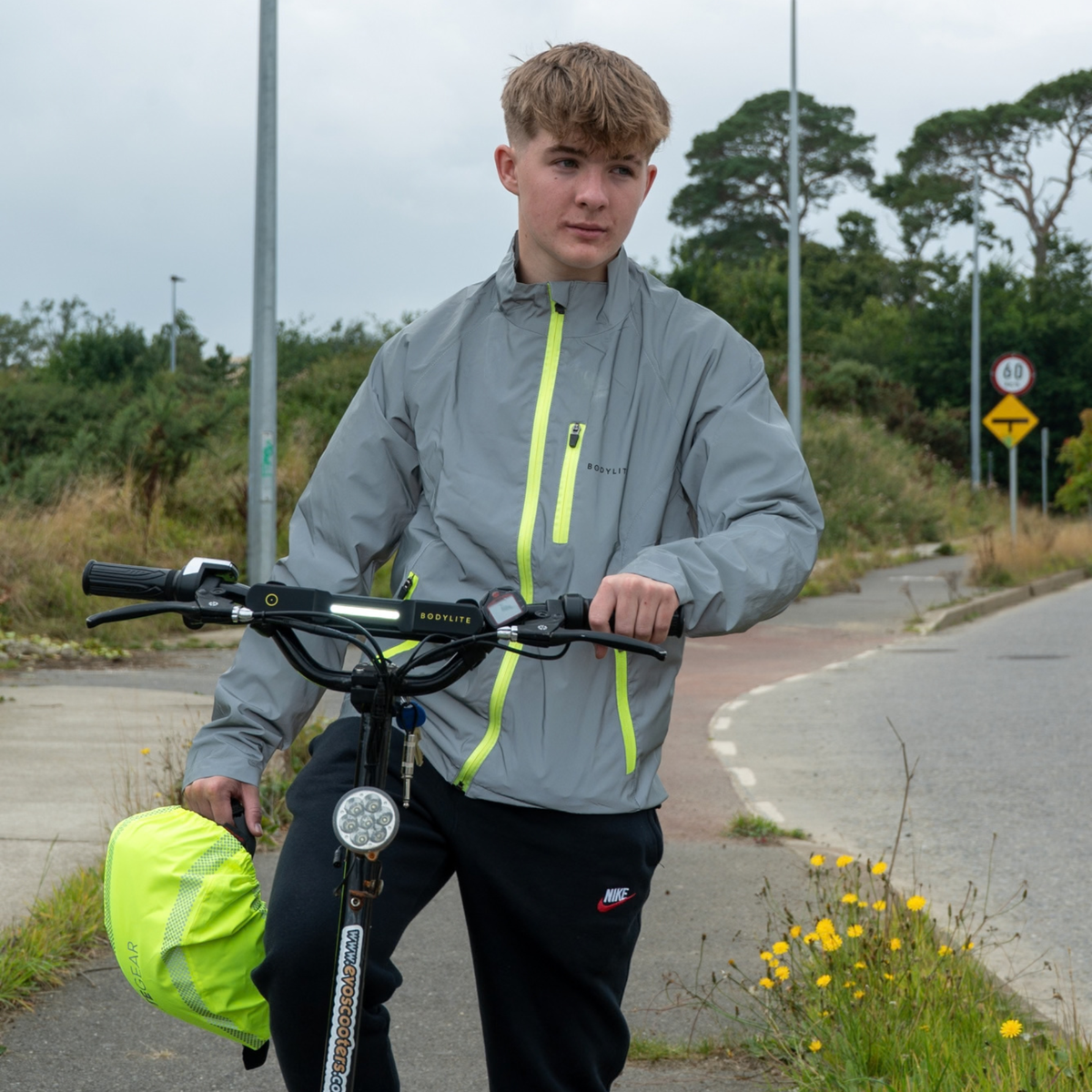 A young man wearing a Bodylite Unisex Fully Reflective Jacket 01303-ALL SIZES stands on a paved road holding a black electric bike. He is also wearing black pants and holding a bright yellow-green helmet.