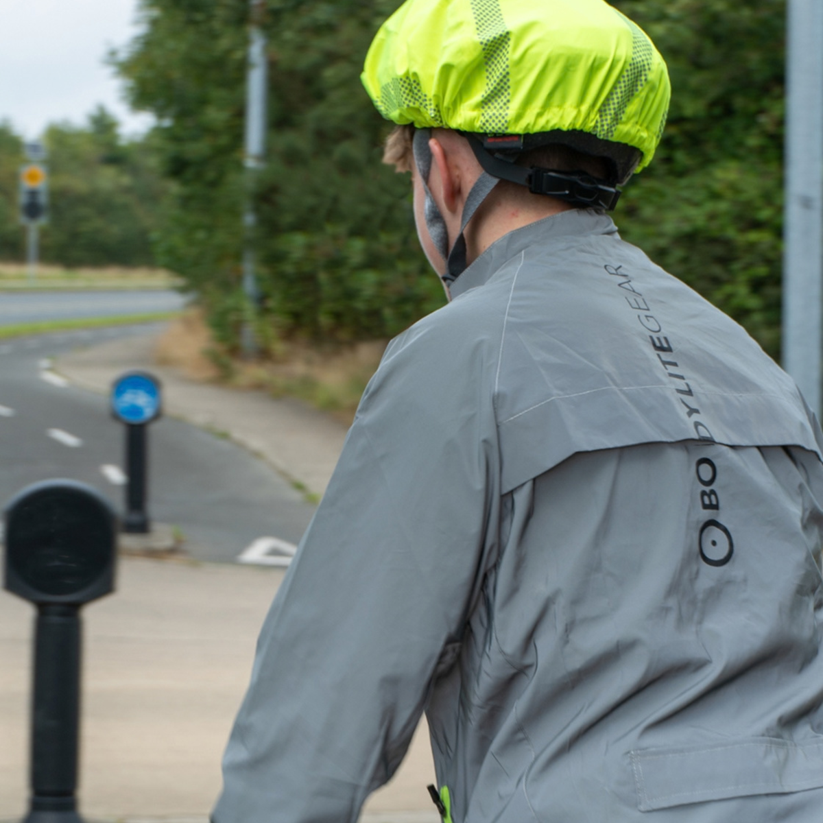 A cyclist viewed from behind wearing a Bodylite Unisex Fully Reflective Jacket 01303-ALL SIZES and a bright neon yellow reflective helmet cover, riding on a paved path outdoors.