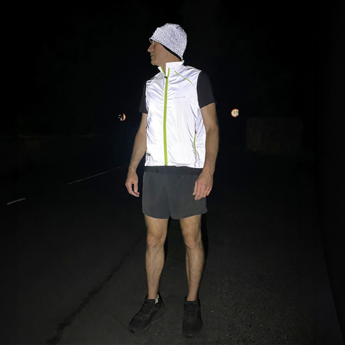 A man wearing a Bodylite Unisex Fully Reflective Vest Gilet 01302-ALL SIZES,  black athletic shorts, and a white reflective Bodylite beanie, standing on a dark road at night.