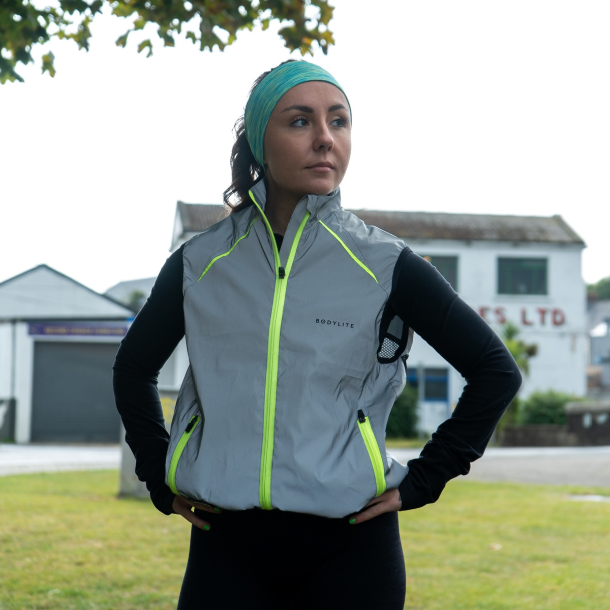 A woman wearing a Bodylite Unisex Fully Reflective Vest Gilet 01302-ALL SIZES and a matching green headband, standing outdoors in front of a white building.