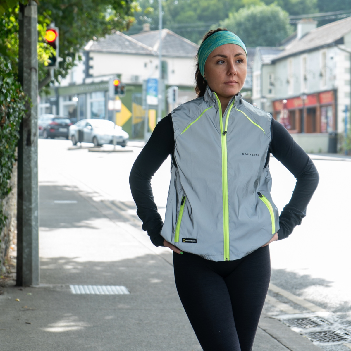 A woman standing on a city street wearing Bodylite Unisex Fully Reflective Vest Gilet 01302-ALL SIZES over a black long-sleeve top and black leggings.