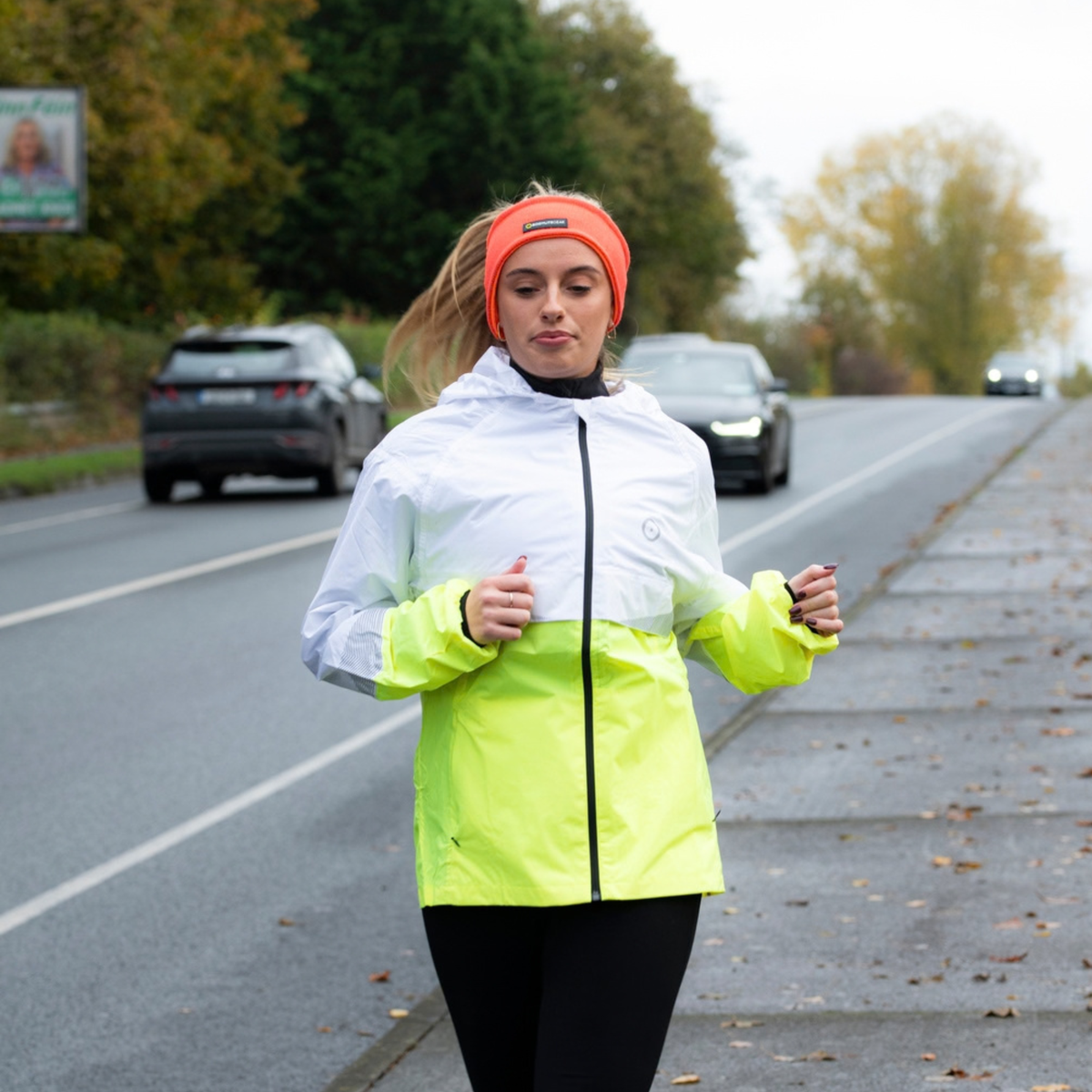A woman jogging outdoors on a road wearing a two-tone reflective jacket with a white upper section and a bright neon yellow-green lower section, paired with a Bodylite Unisex Reflective Headband Neon Orange 01301-ALL SIZES.