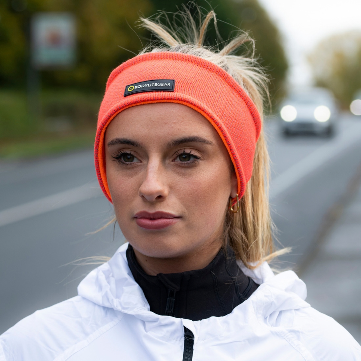 A woman wearing a Bodylite Unisex Reflective Headband Neon Orange 01301-ALL SIZES, photographed outdoors on a blurred road background.