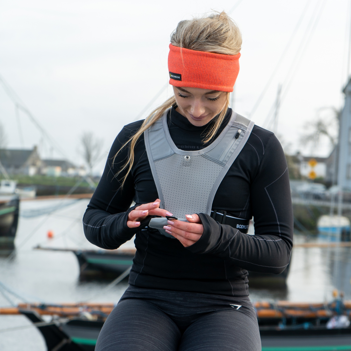 A woman in athletic wear stands by a harbor, looking down at a phone. She is wearing a grey running vest, a black long-sleeve top, black leggings, and a Bodylite Unisex Reflective Headband Neon Orange 01301-ALL SIZES.
