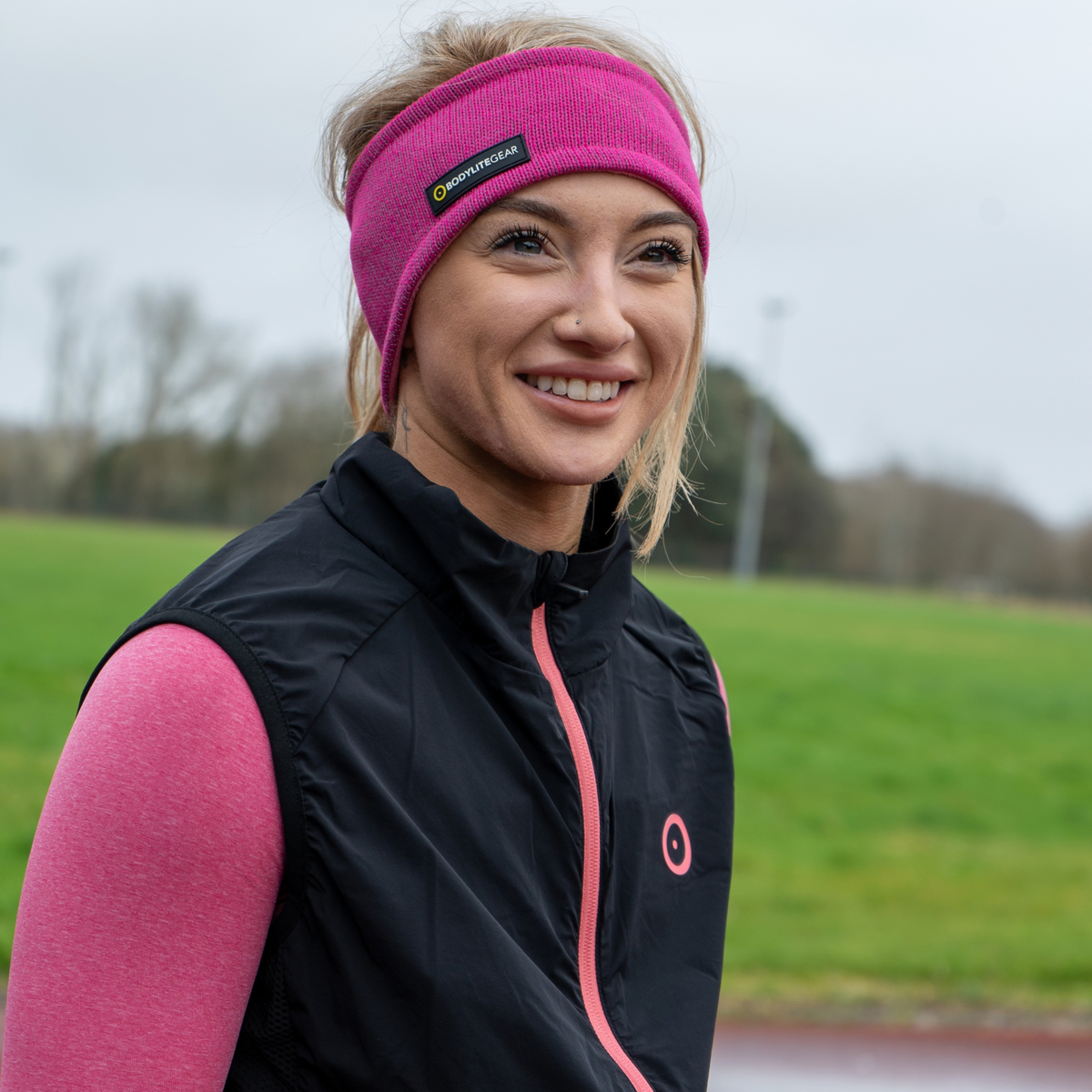 A smiling woman outdoors wearing a Bodylite Unisex Reflective Headband Neon Pink 01301-ALL SIZES, paired with a black sleeveless zip vest over a pink long-sleeve top.