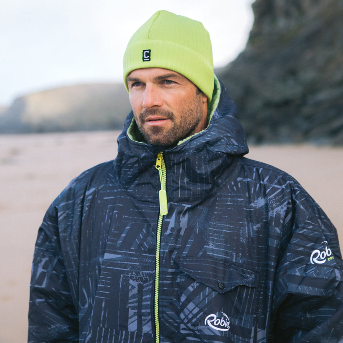 Man wearing a C-Skins Unisex Storm Chaser Thermal Neoprene Swim Watersports Beanie Lime C-HOBE and a hardshell changing robe on the beach