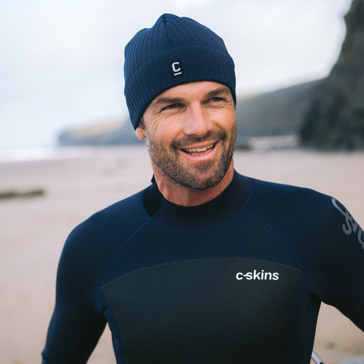 Man wearing a C-Skins Unisex Storm Chaser Thermal Neoprene Swim Watersports Beanie Slate Navy C-HOBE and a wetsuit on a beach