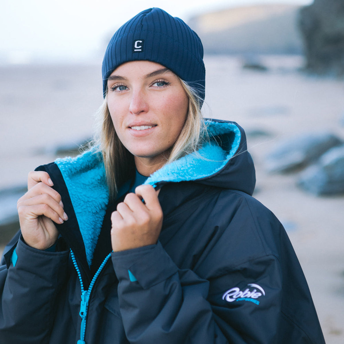 Woman wearing a C-Skins Unisex Storm Chaser Thermal Neoprene Swim Watersports Beanie Slate Navy C-HOBE and a hardshell changing robe on a beach