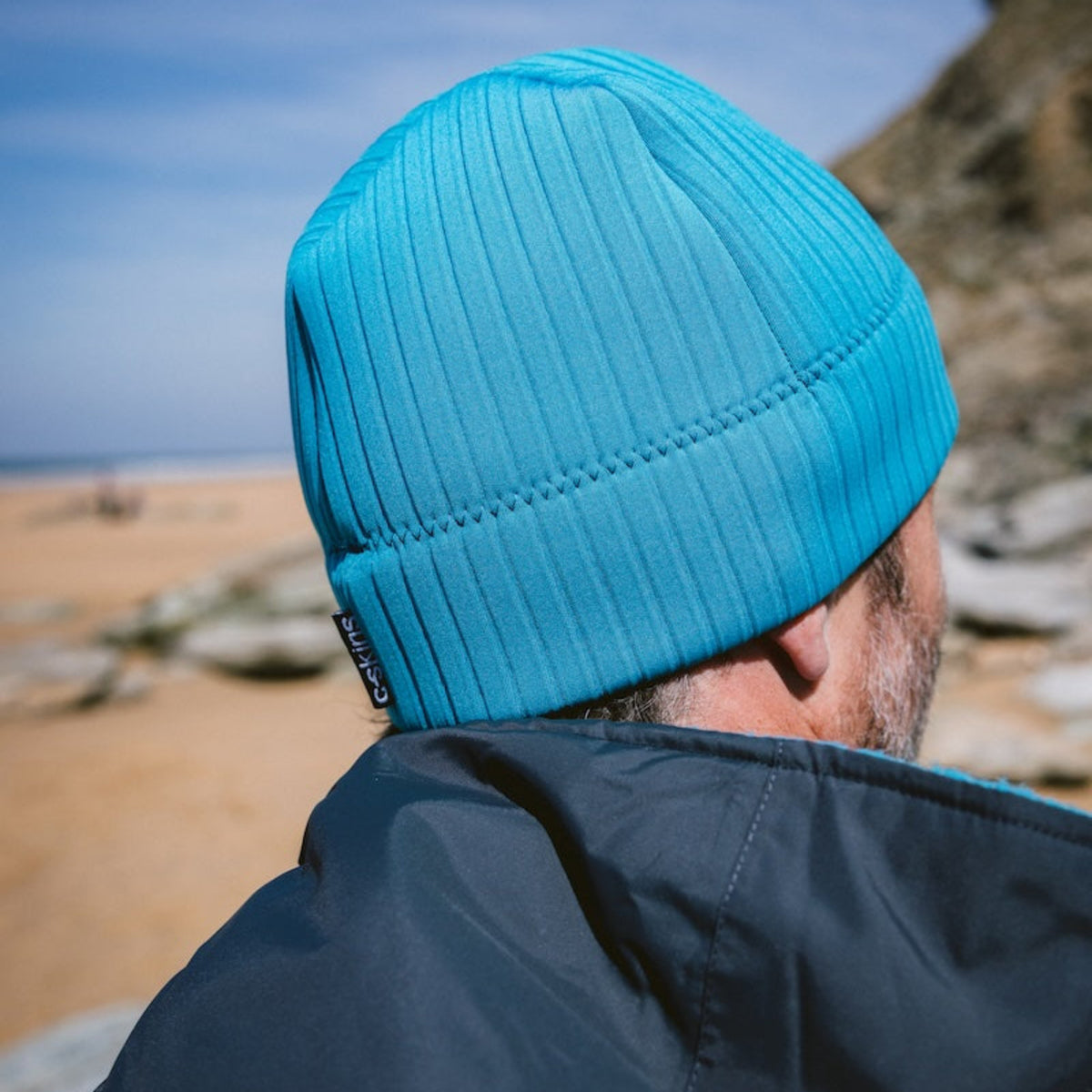 Man wearing a black hardshell changing robe with blue accents and a C-Skins Unisex Storm Chaser Thermal Neoprene Swim Watersports Beanie Teal Blue C-HOBE on a beach