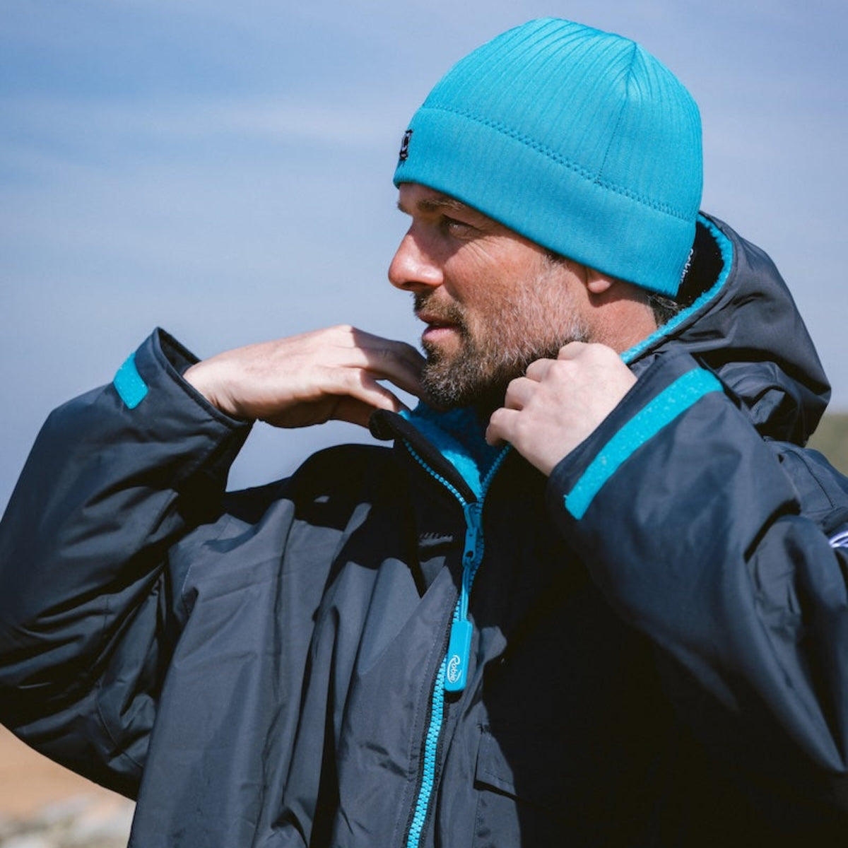 Man wearing a black hardshell changing robe with blue accents and a C-Skins Unisex Storm Chaser Thermal Neoprene Swim Watersports Beanie Teal Blue C-HOBE on a beach