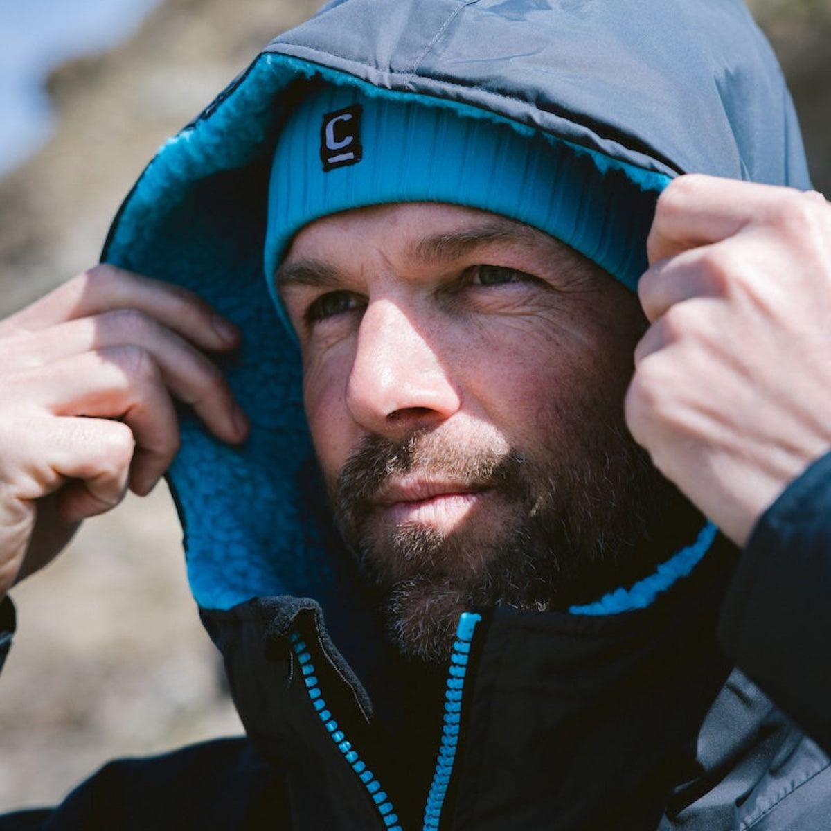 Man wearing a black hardshell changing robe with blue accents, with the hood up, and a C-Skins Unisex Storm Chaser Thermal Neoprene Swim Watersports Beanie Teal Blue C-HOBE on a beach