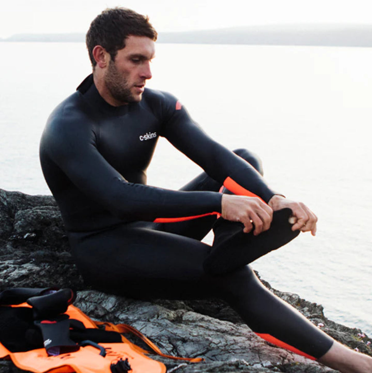 Man wearing C-Skins Unisex Freedom Swim 4mm GBS Neoprene Thermal Swim Watersports Socks C-SOXSR4 and a wetsuit on a rocky outcrop beside the sea