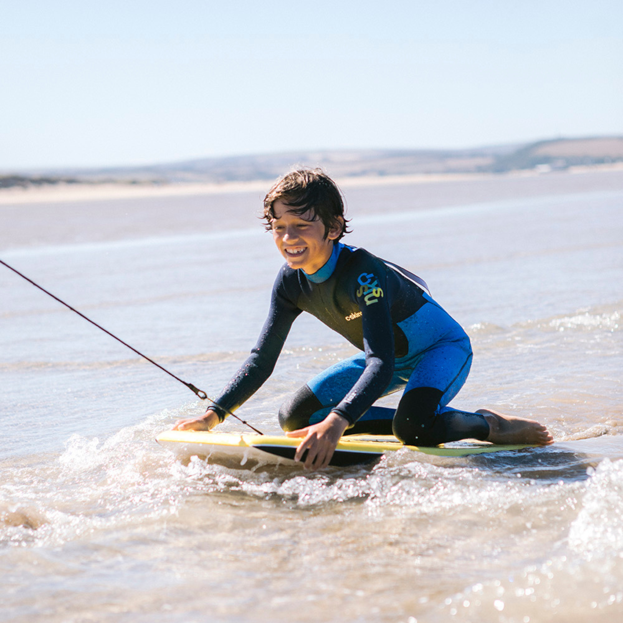 Kid wearing C-Skins Junior Unisex Element 3mm 2mm Flatlock Steamer Wetsuit Cyan Slate Multi C-EL32STJ getting pulled along on a bodyboard on the beach