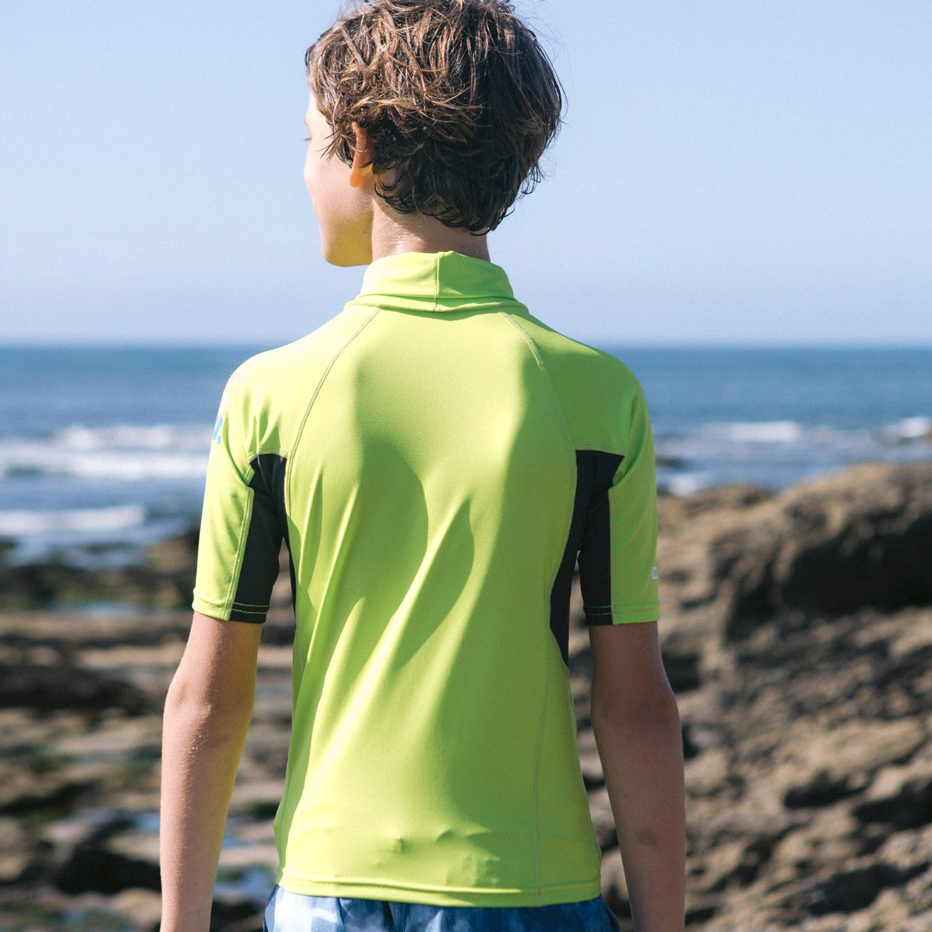 Young boy wearing C-Skins Junior Kids NuWave Rash X UPF50+ Short Sleeve Rash Vest Lime Anthracite Multi C-NLYSSJ with a blurry beach background. Back of rash vest being shown