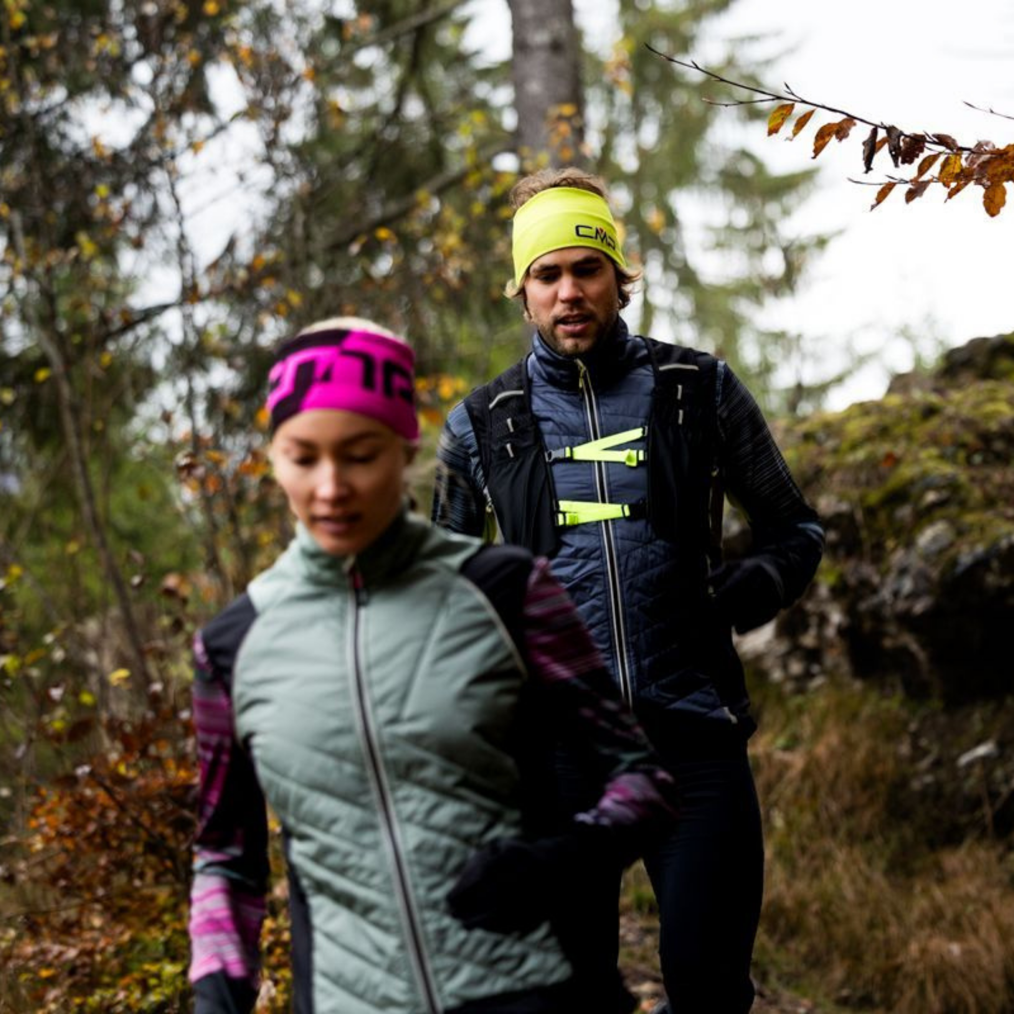 Two trail runners jogging on an outdoor forest path. The runner in the foreground wears a light sage green puffer vest over a dark long-sleeve shirt, black gloves, and a bright pink and black headband. The man running behind her wears a CMP Unisex Santa Fe Trail Running Vest Titanio U911 3V31537