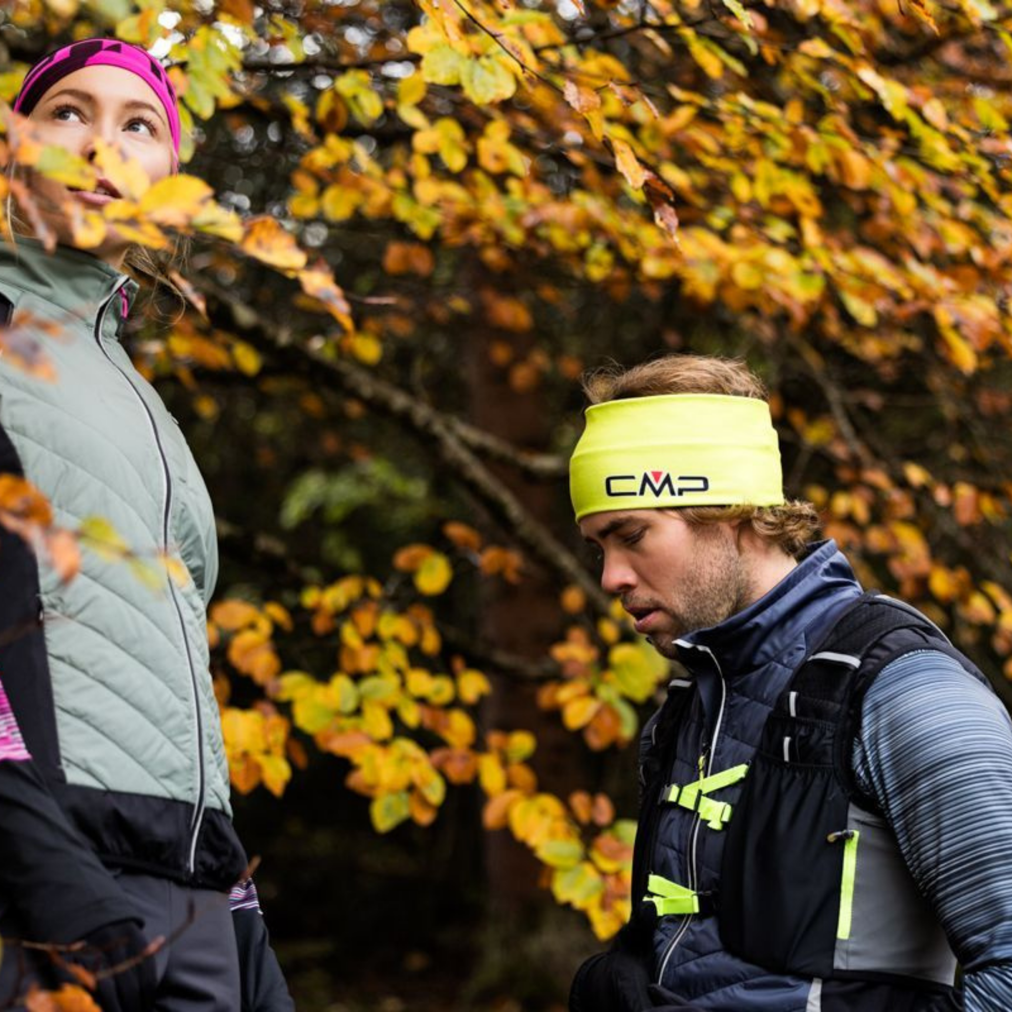 Two trail runners outdoors in autumn foliage, with the man wearing a bright yellow CMP headband and a CMP Unisex Santa Fe Trail Running Vest Titanio U911 3V31537.