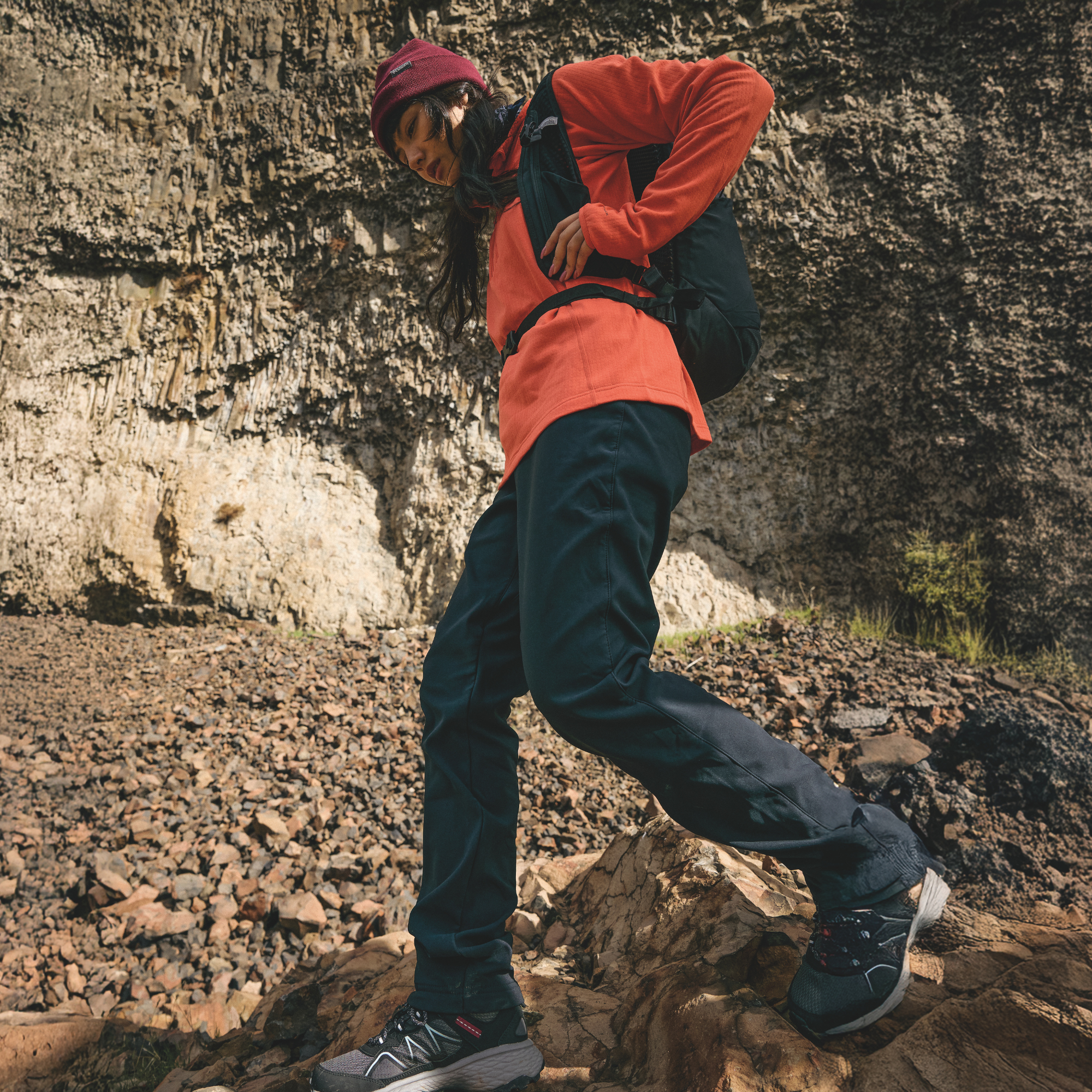 A person hiking on a rocky cliffside path, wearing a bright orange long-sleeve top, dark navy pants, a burgundy beanie, and a Columbia Blackcomb Ridge 18L Backpack Black 010 2121411