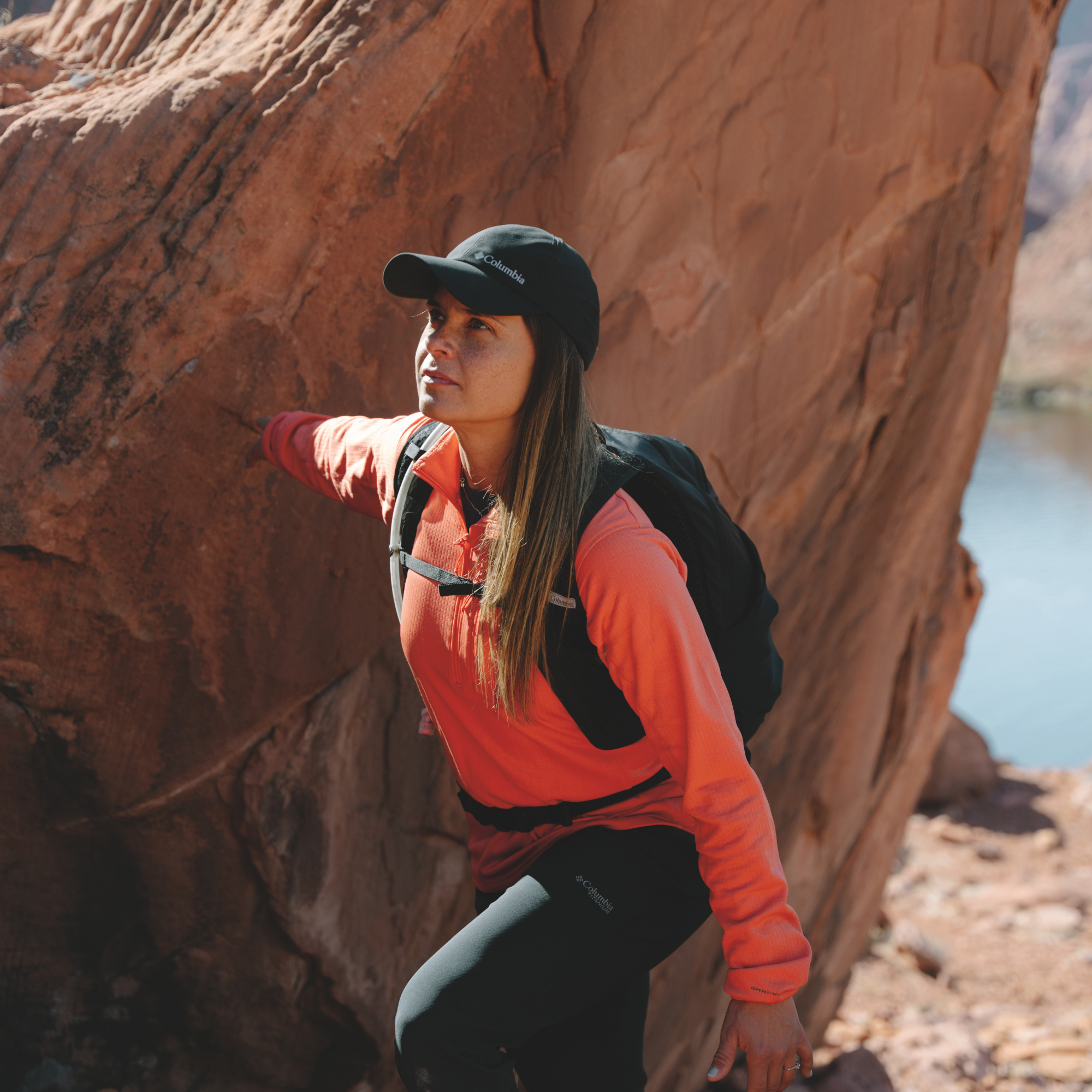 A woman hiking outdoors against a large red rock formation, wearing a bright coral-orange long-sleeve top, black trousers, a black cap, and a Columbia Blackcomb Ridge 18L Backpack Black 010 2121411