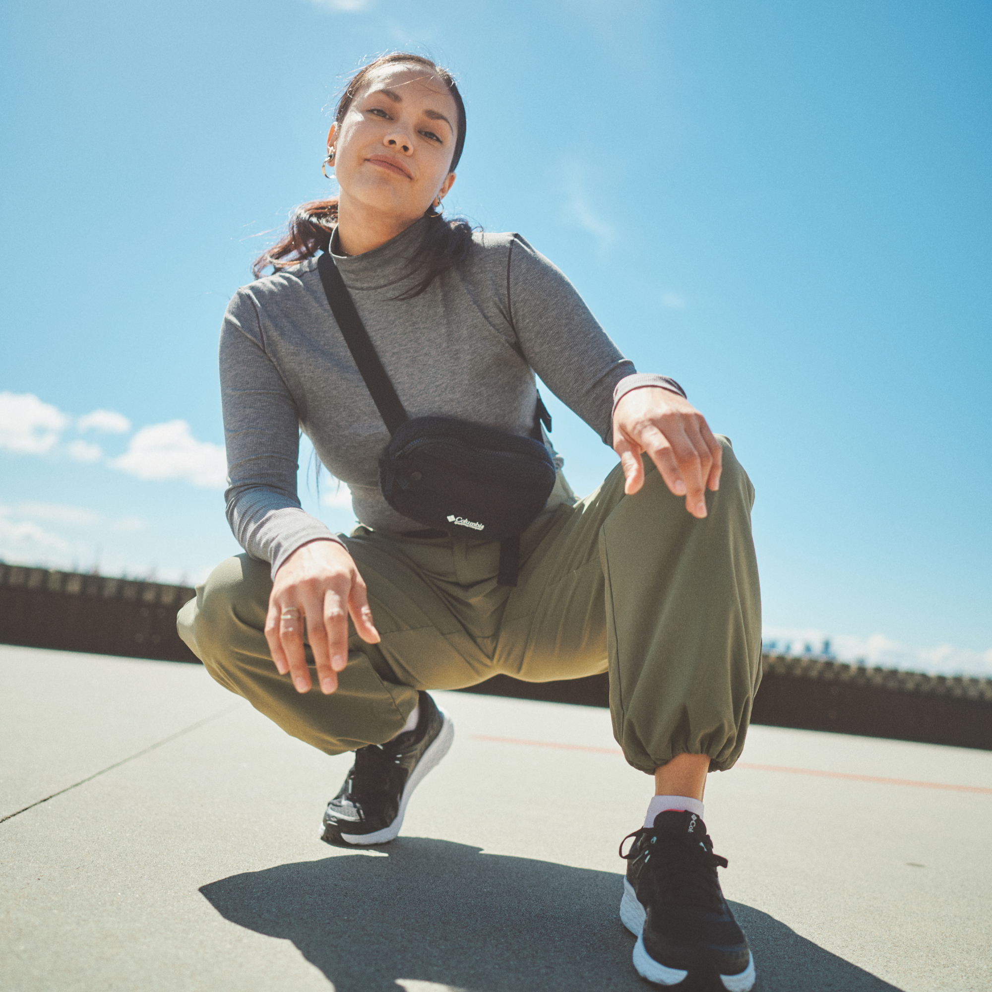 A woman crouches outdoors under a bright blue sky wearing a grey turtleneck, olive green jogger pants, black and white sneakers, and a Columbia Helvetia II Sherpa Hip Pack Black 010 2094541