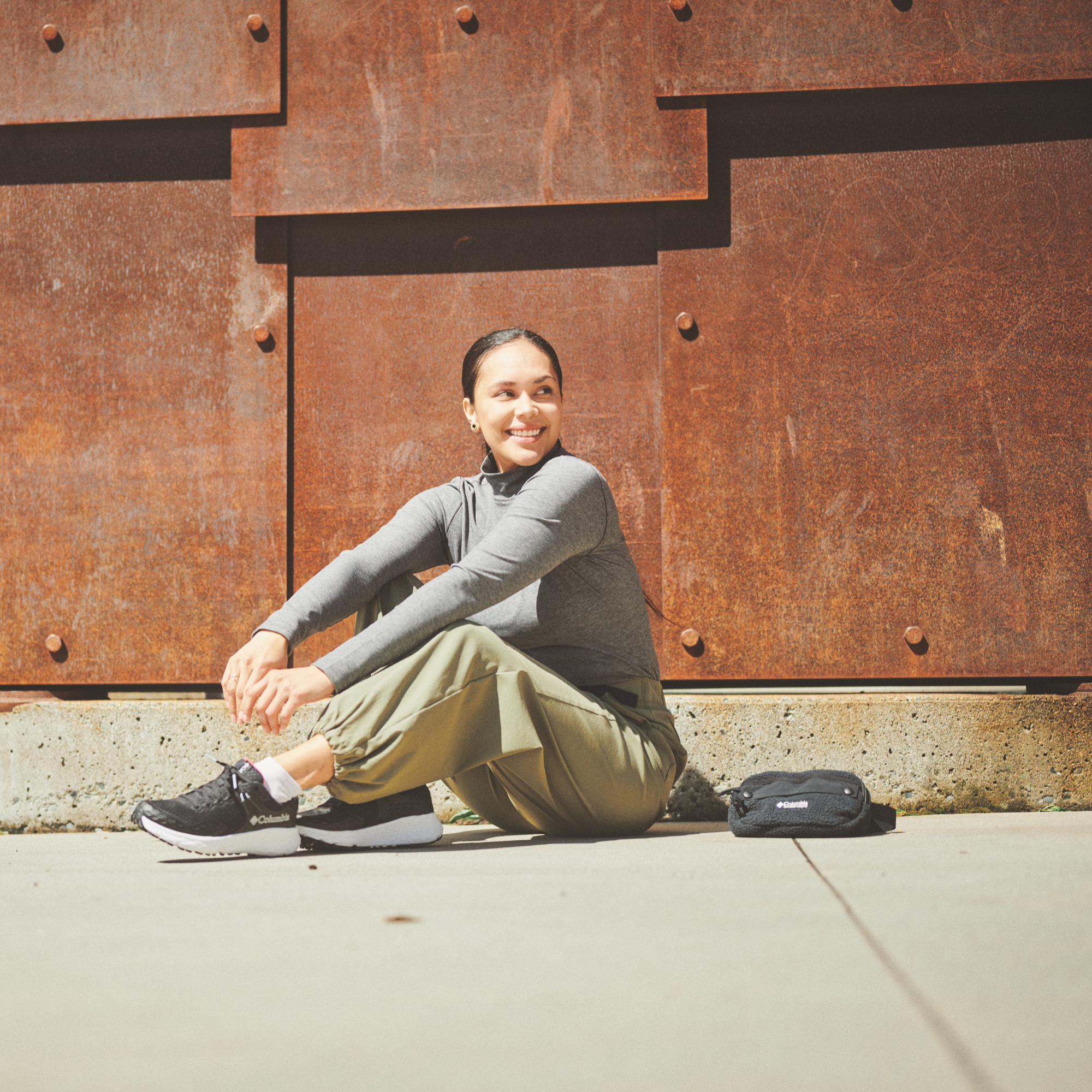 A woman sits on the ground in front of a large rusted metal wall, wearing a grey long-sleeve athletic top, olive green jogger pants, and black running shoes. A Columbia Helvetia II Sherpa Hip Pack Black 010 2094541 rests on the ground beside her.