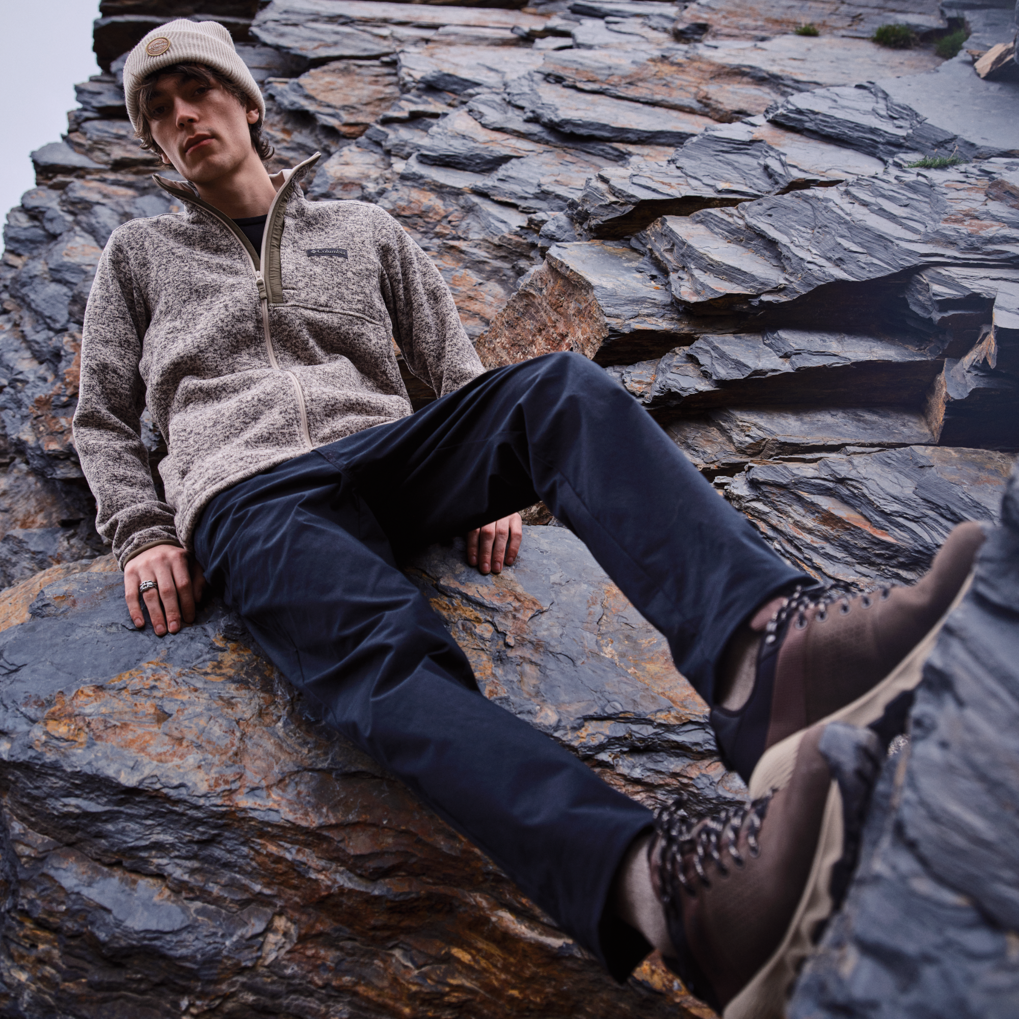 A man reclining on a large rock wearing a beige fleece zip-up jacket, dark navy pants, beige cap and Columbia Men's Newton Nimble LTR Waterproof Trail Shoes Tundra Black 357 2140371