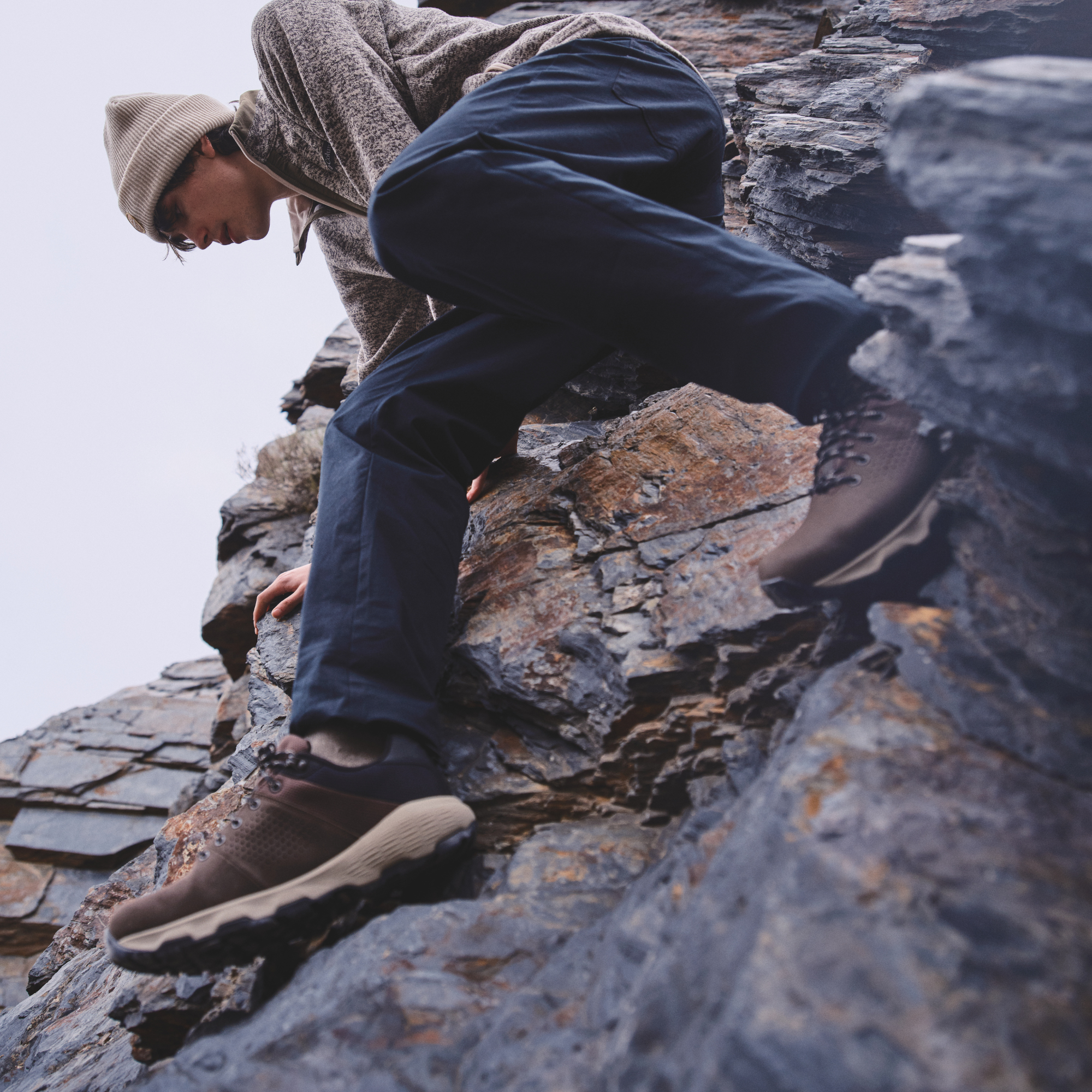 A person climbing a rocky outdoor cliff, wearing navy trousers, a beige beanie, and a textured knit sweater and Columbia Men's Newton Nimble LTR Waterproof Trail Shoes Tundra Black 357 2140371 against a pale sky.