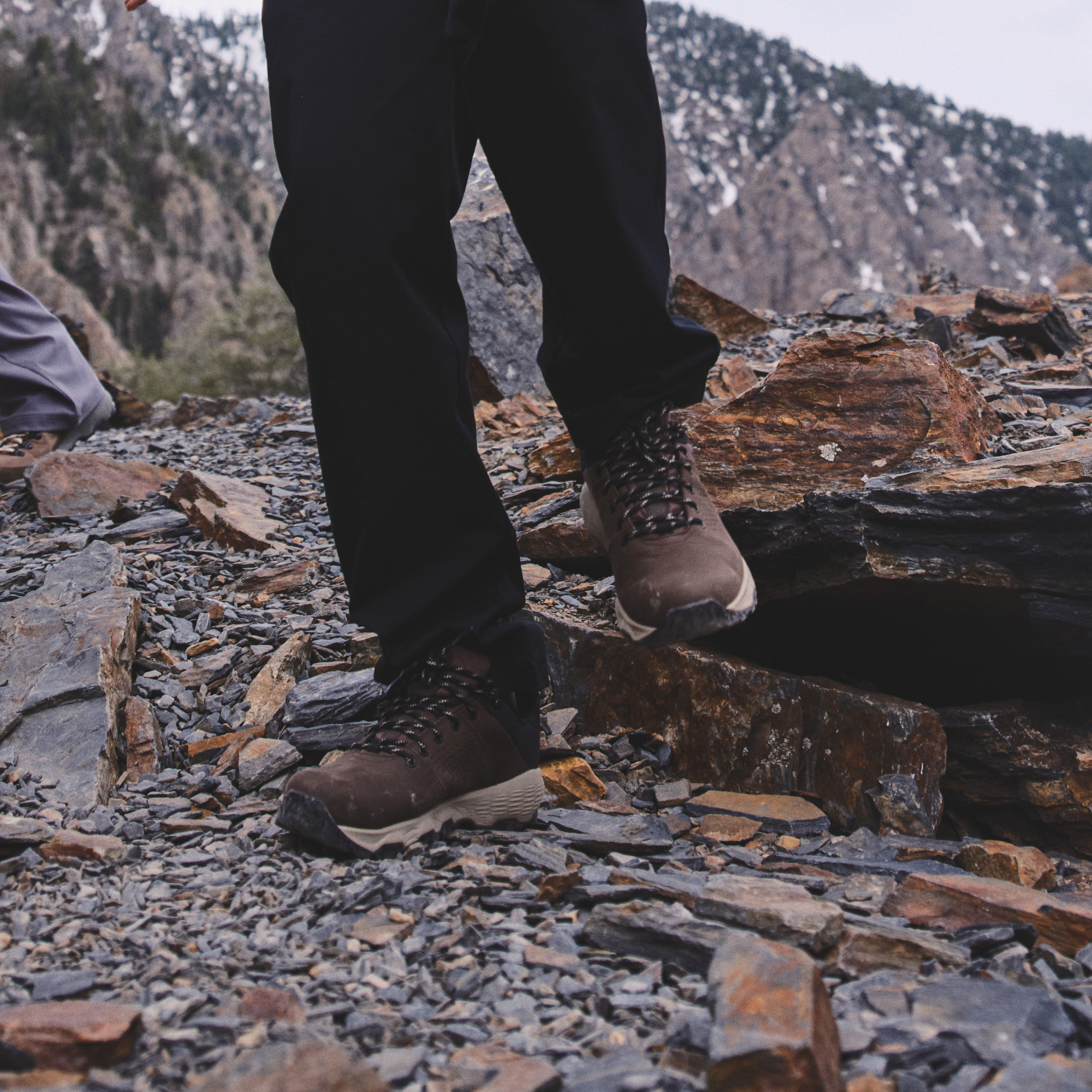 A person wearing black pants and Columbia Men's Newton Nimble LTR Waterproof Trail Shoes Tundra Black 357 2140371 walking on a rocky, gravel-covered outdoor trail with mountains in the background.