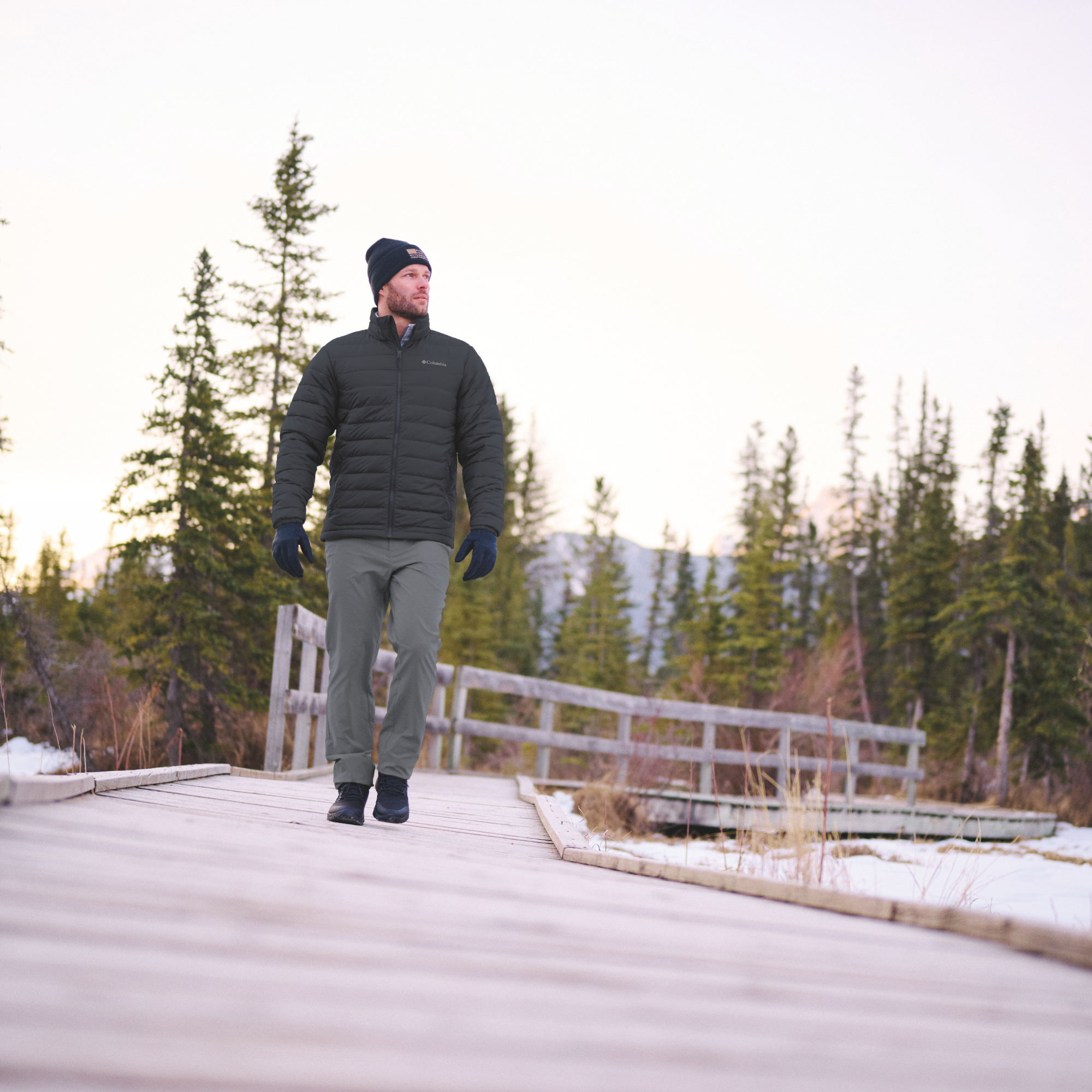 A man wearing a Columbia Men's Powder Lite II Insulated Jacket Black 010 2086964, dark beanie, black gloves, and grey trousers walks along a wooden boardwalk surrounded by evergreen trees and light snow.