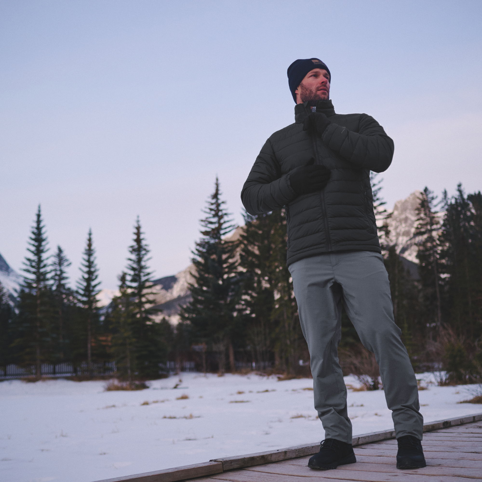A man wearing a Columbia Men's Powder Lite II Insulated Jacket Black 010 2086964, grey trousers, and a black beanie stands on a wooden boardwalk in a snowy mountain setting.
