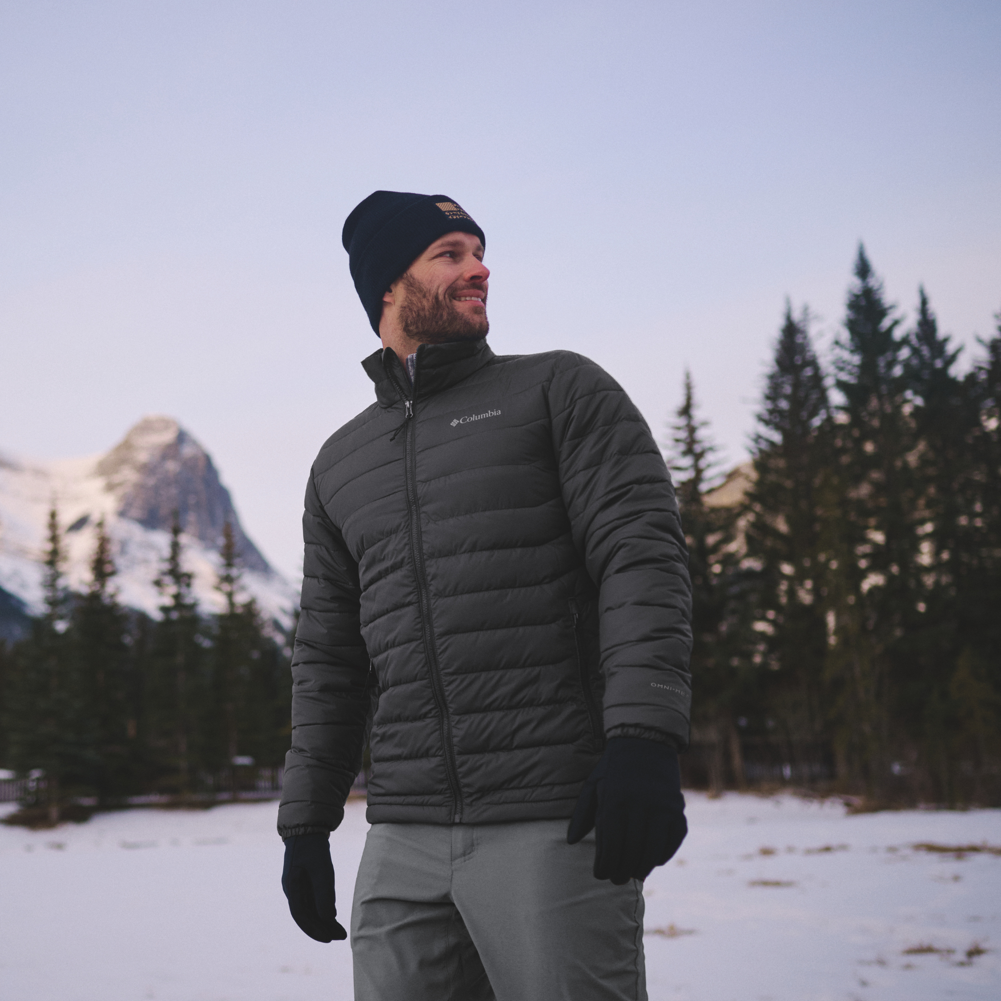 A man wearing a Columbia Men's Powder Lite II Insulated Jacket Black 010 2086964, black beanie, and black gloves stands in a snowy outdoor setting with pine trees and mountains in the background.