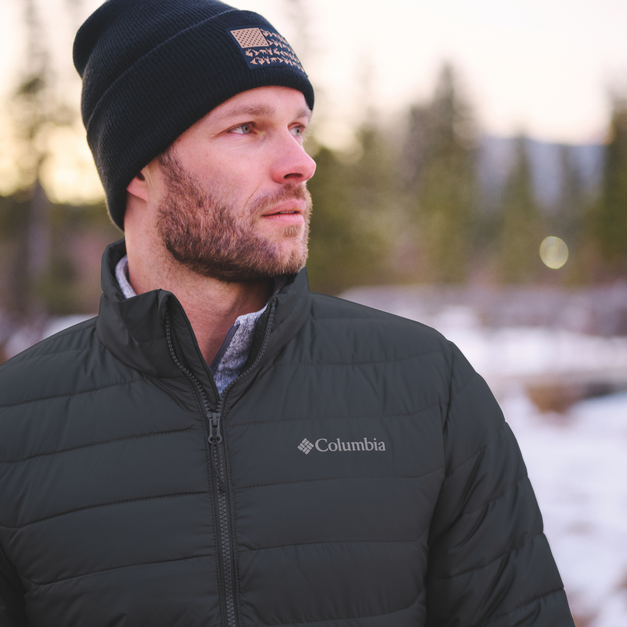 A man wearing a Columbia Men's Powder Lite II Insulated Jacket Black 010 2086964 and a black beanie with a logo patch, photographed outdoors in a snowy, blurred winter setting.