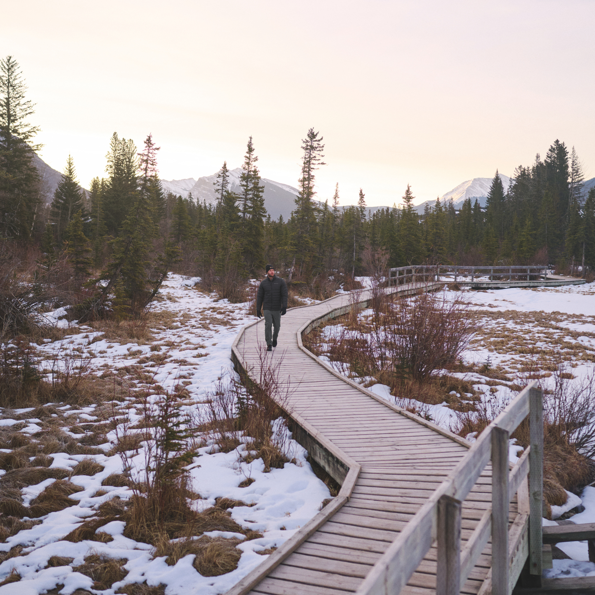 A person walks along a curved wooden boardwalk through a snowy landscape with evergreen trees and distant mountains at sunset wearing a Columbia Men's Powder Lite II Insulated Jacket Black 010 2086964.