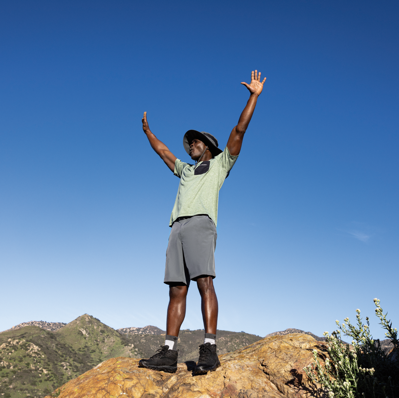 Person standing on a rocky outcrop with arms raised against a clear blue sky wearing Columbia Unisex Bora Bora UPF50 Booney Sun Hat Grill 028 1447091