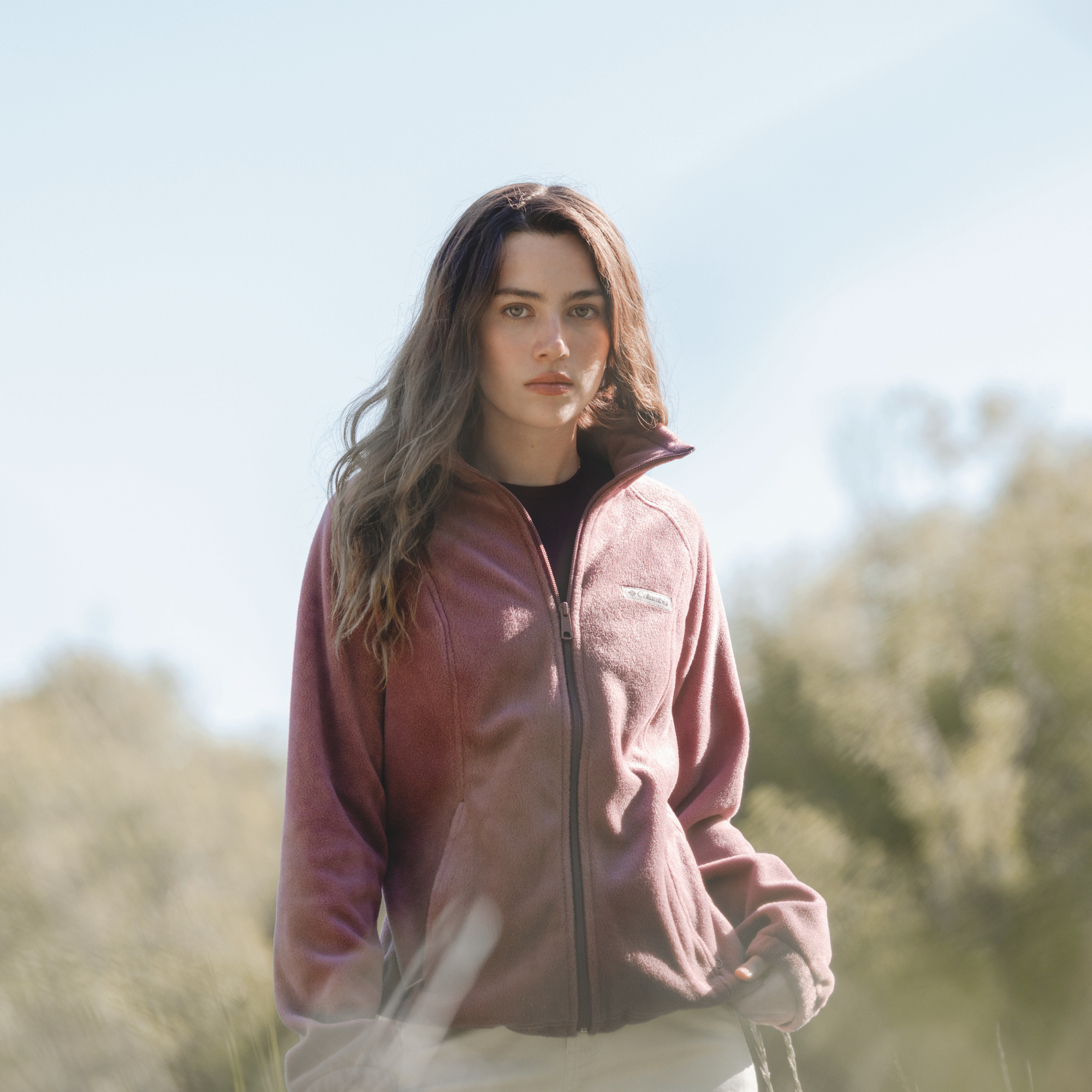 A woman wearing a Columbia Women's Benton Springs Full Zip Fleece Jacket Antique Mauve 553 1372111 stands outdoors in a grassy field with a soft-focus natural background.