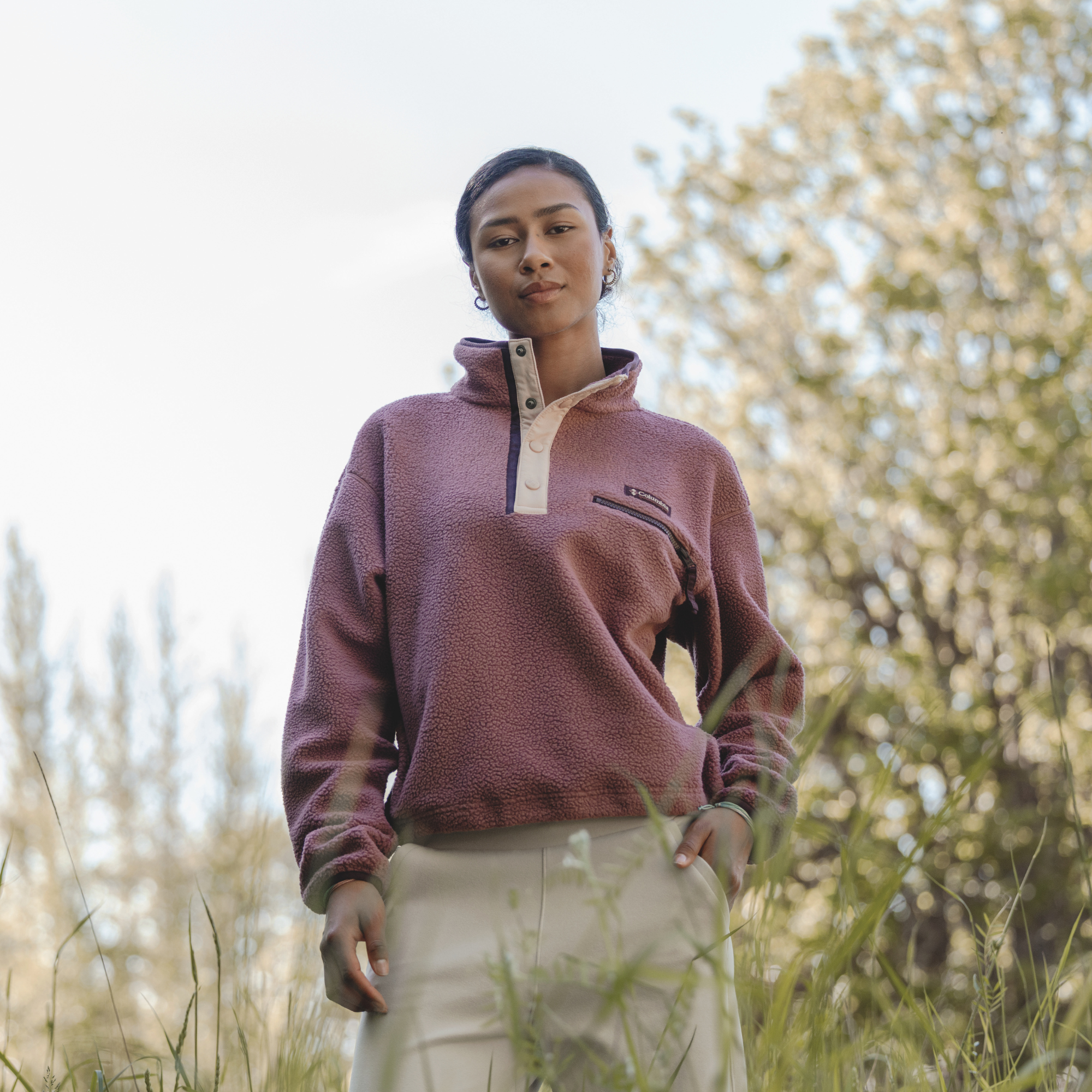 A woman wearing a Columbia Women's Helvetia II Cropped Sherpa Half Snap Fleece Antique Mauve Mineral Pink 552 2092231 standing outdoors in a grassy field with trees in the background.