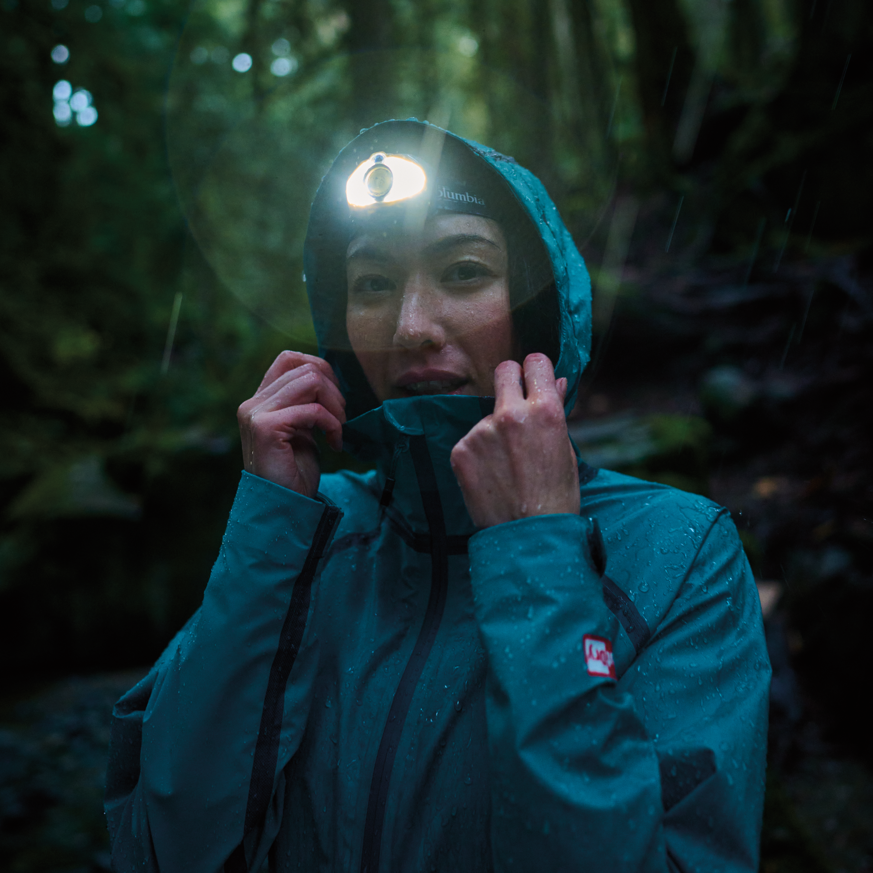 Woman hiking in a dark rainy forest, wearing a Columbia Women's Reign No Shine Outdry Extreme Waterproof Jacket River Blue 364 2116293 with the hood up and a headtorch