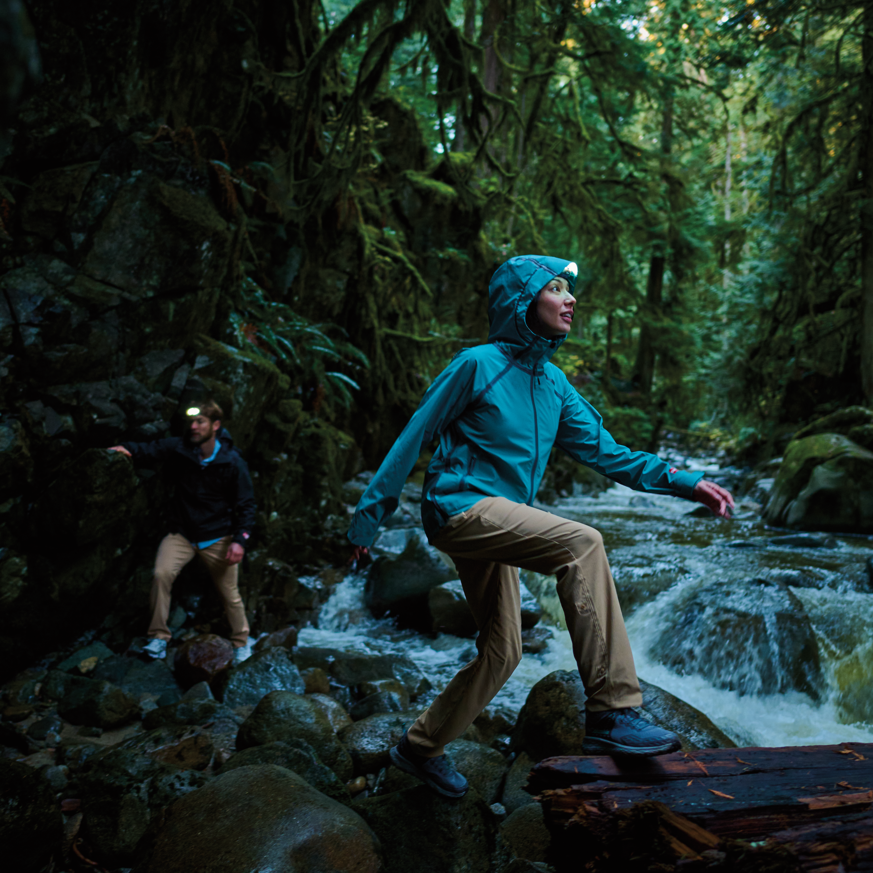 Woman hiking in a dark forest, by a river, wearing Columbia Women's Reign No Shine Outdry Extreme Waterproof Jacket River Blue 364 2116293 and a headtorch