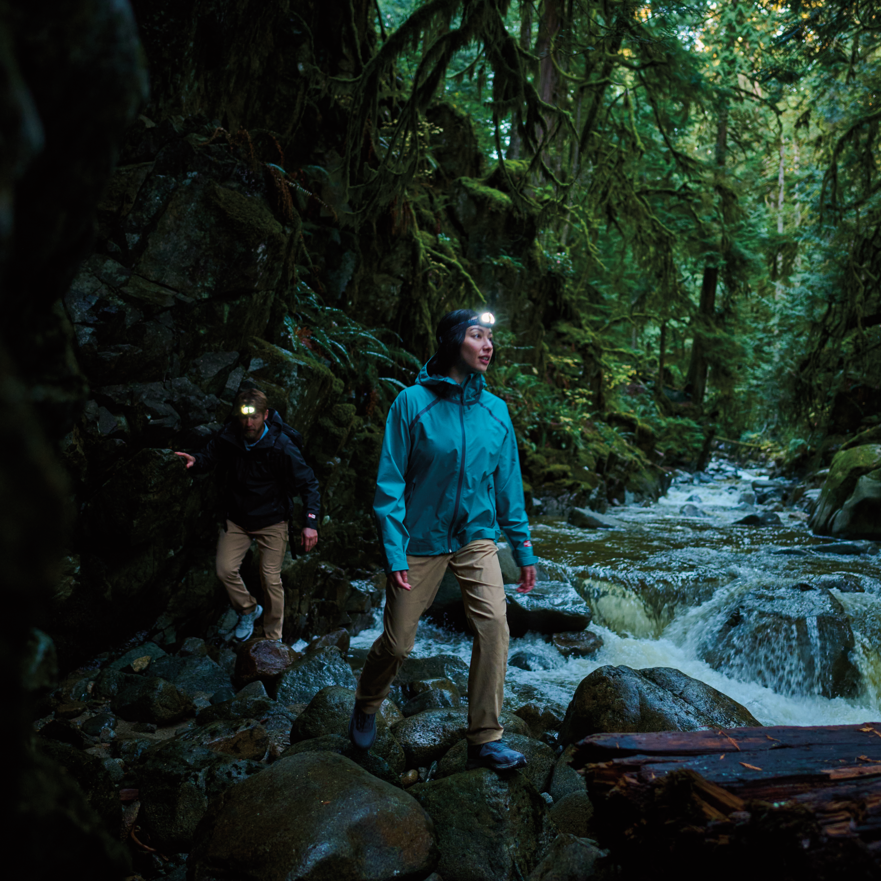 Two hikers hiking in a dark forest beside a river. Woman wearing Columbia Women's Reign No Shine Outdry Extreme Waterproof Jacket River Blue 364 2116293 and a headtorch