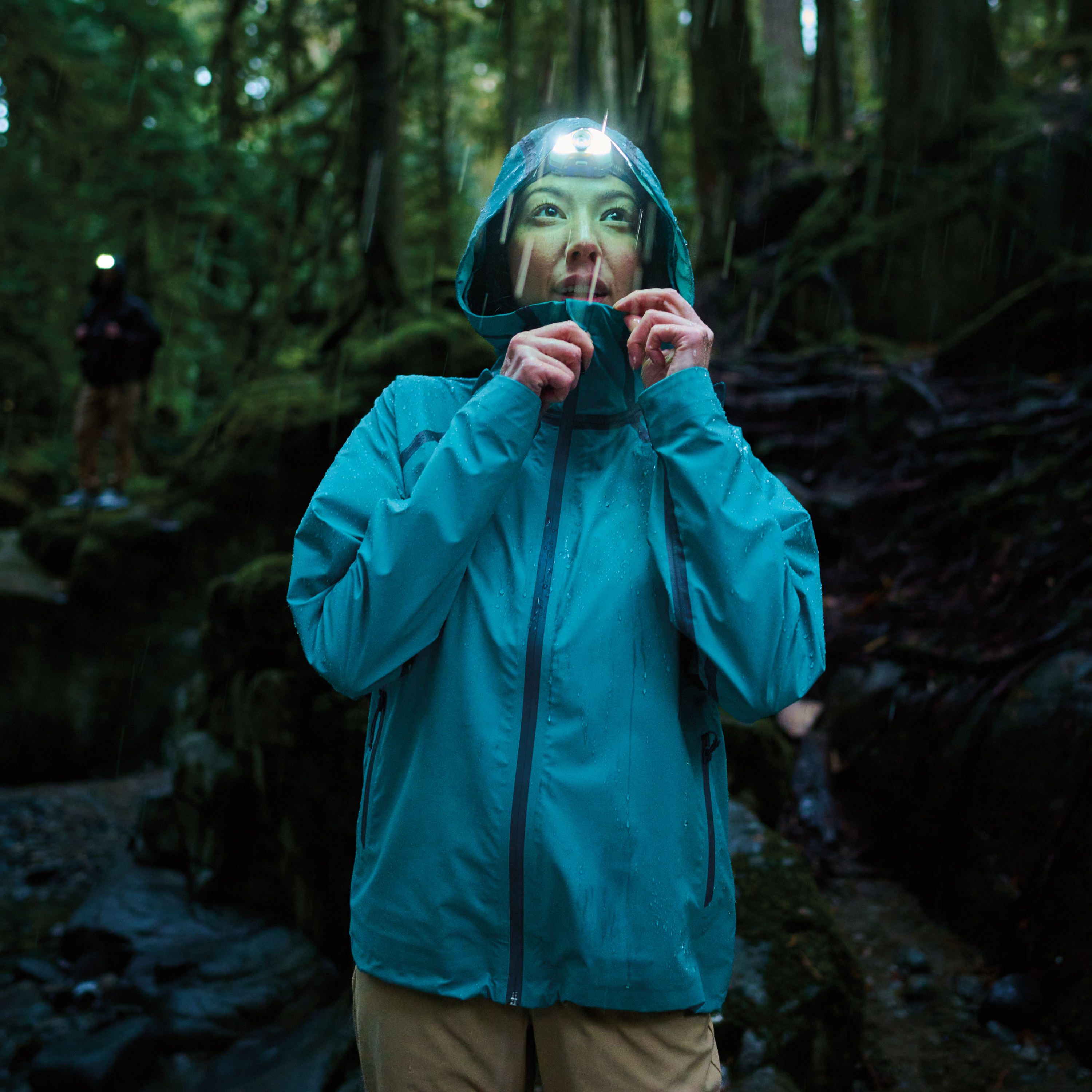 Woman standing in a dark rainy forest with her hood up and headtorch on her head. Wearing Columbia Women's Reign No Shine Outdry Extreme Waterproof Jacket River Blue 364 2116293.