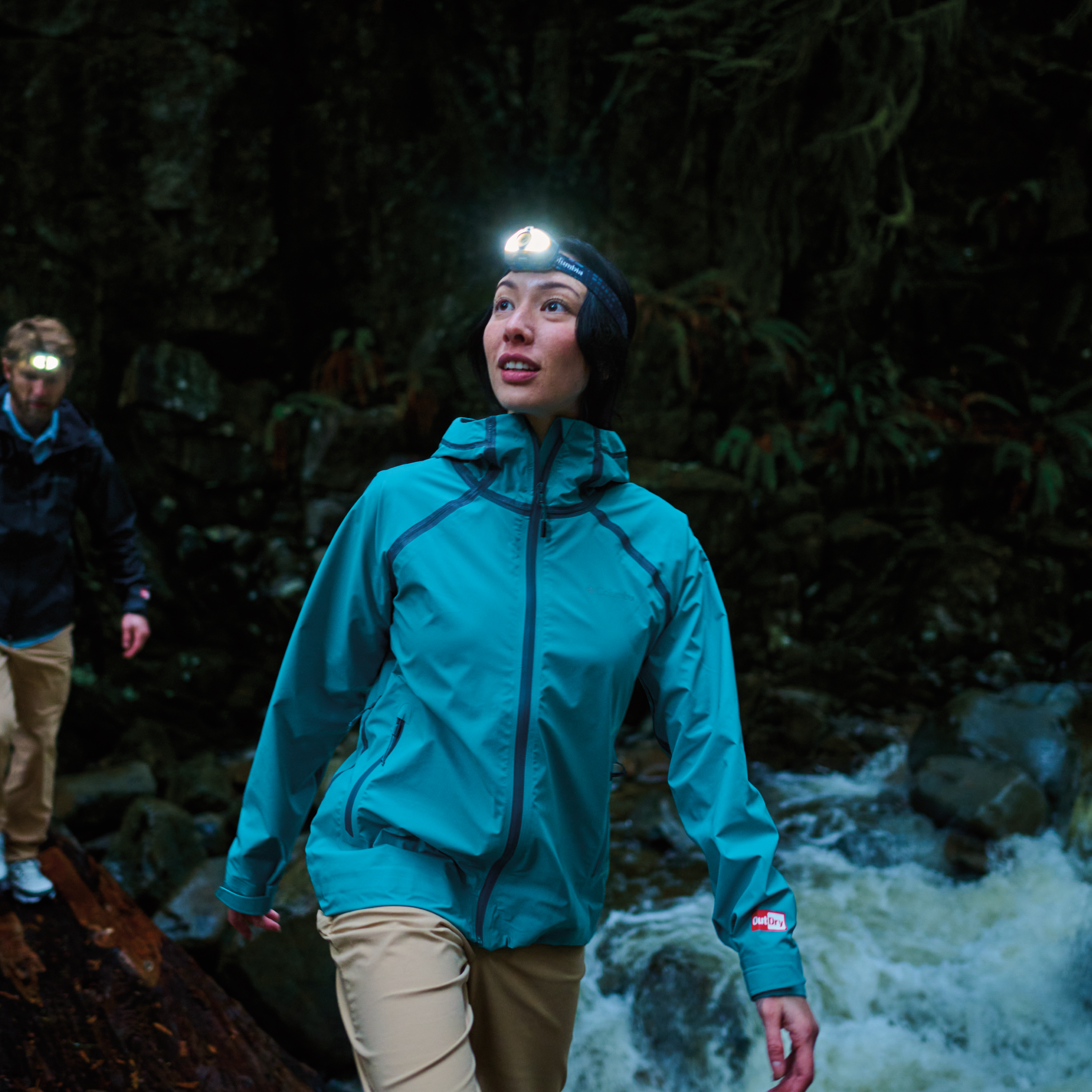 Woman hiking in a dark forest by a river, wearing Columbia Women's Reign No Shine Outdry Extreme Waterproof Jacket River Blue 364 2116293 and a headtorch