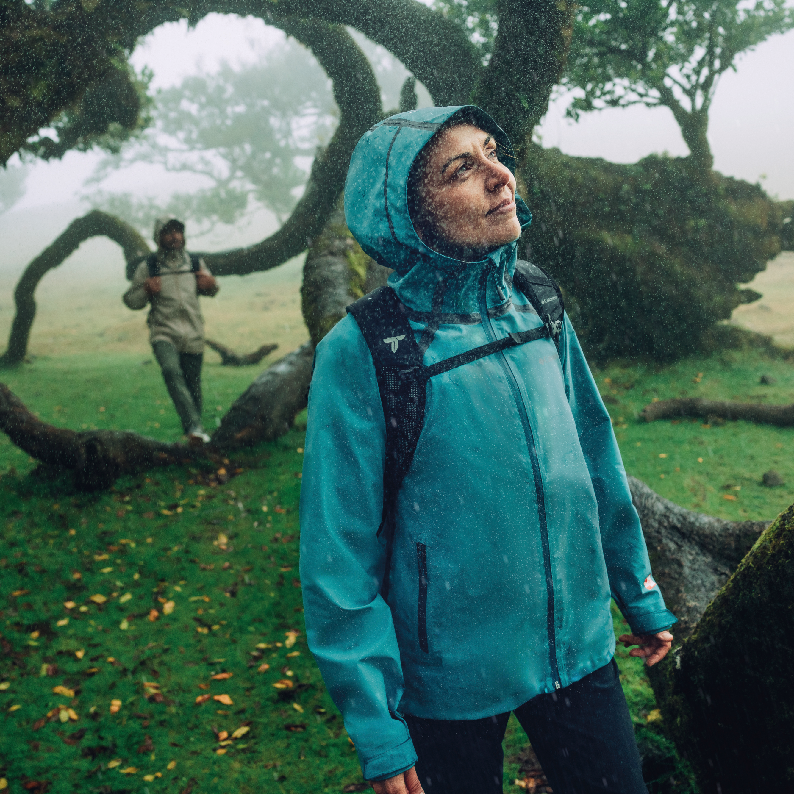 Woman wearing a Columbia Women's Reign No Shine Outdry Extreme Waterproof Jacket River Blue 364 2116293 standing in a foggy, forested area with another person in the background.