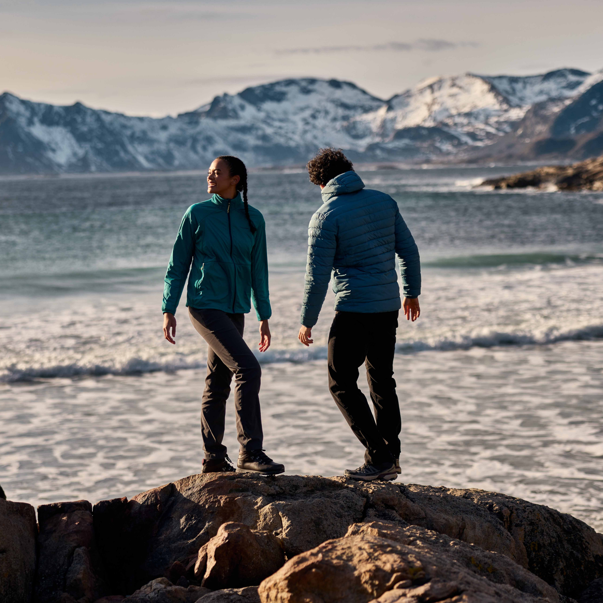 Two people standing on rocks by the ocean, wearing outdoor jackets and trousers, with snowy mountains in the background. Man wears Craghoppers Men's Complite IX Hooded Insulated Jacket Winter Storm 1Y0 CMN323