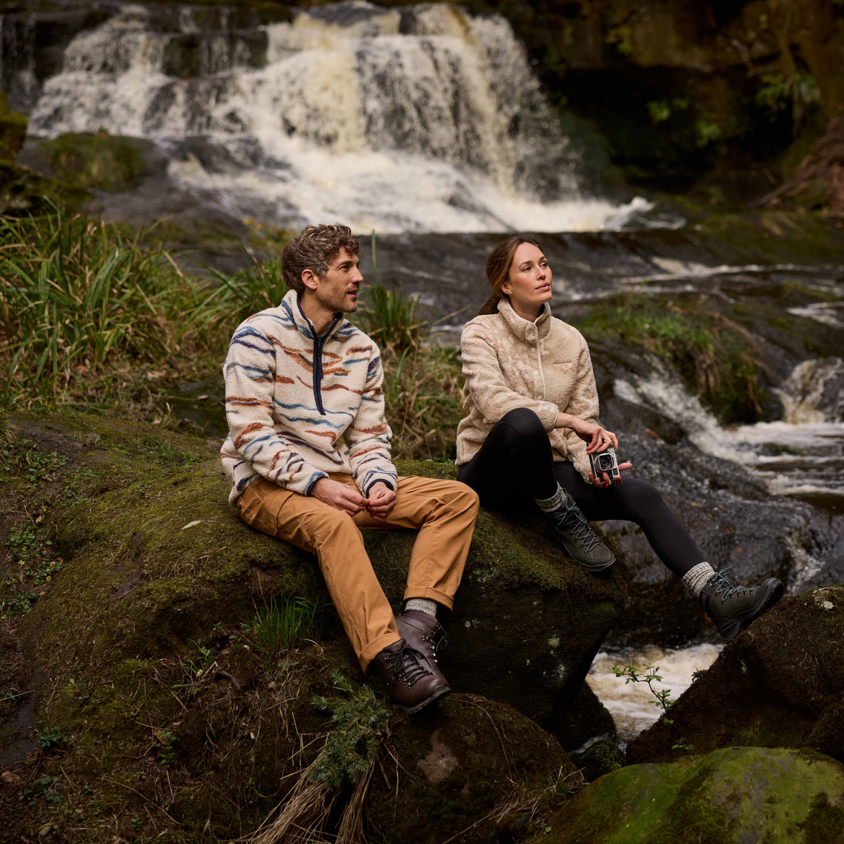 Man wearing Craghoppers Men's Rawthey National Trust Half Zip Fleece Top Ecru Winterstorm 1XJ CMA1425 sitting on mossy groudn beside a river with a woman in a sherpa fleece sitting beside him