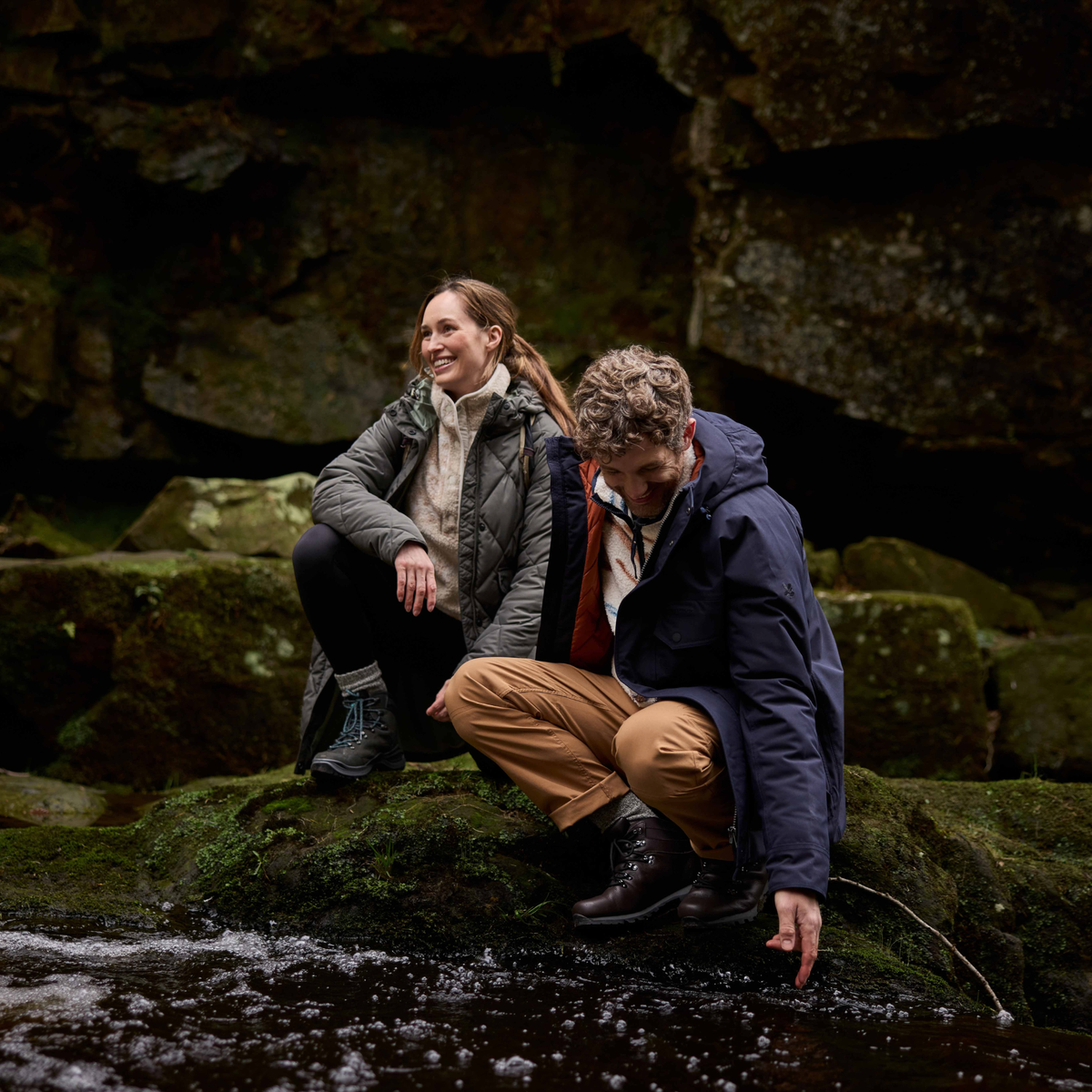 Man wearing Craghoppers Men's Rawthey National Trust Half Zip Fleece Top Ecru Winterstorm 1XJ CMA1425 and a navy waterproof jacket leaning down to put his hand in a river