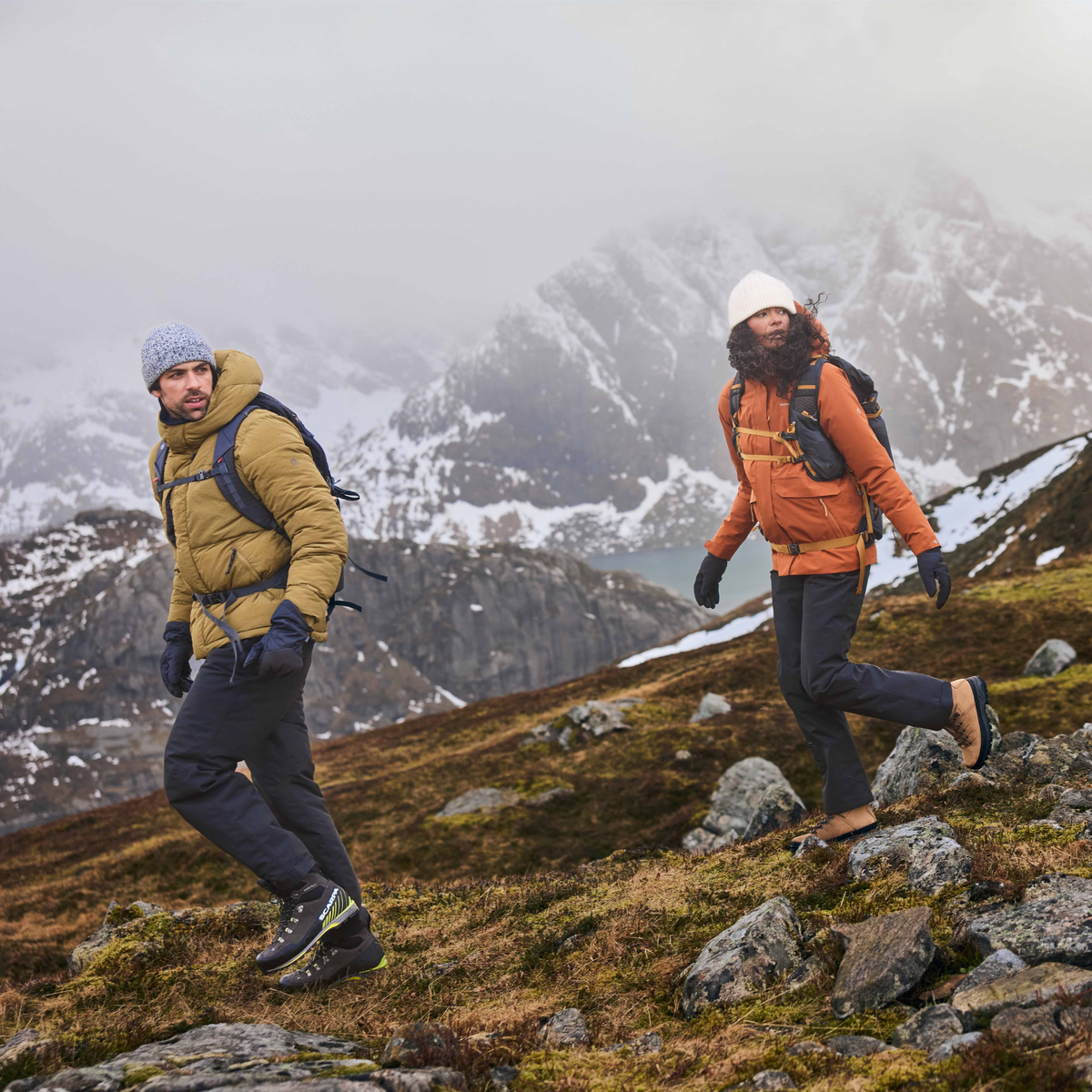 Two hikers wearing outdoor winter jackets and backpacks traverse a rocky, grassy mountain slope with snowy peaks in the background under a foggy sky. Man wearing Craghoppers Men's Steall Pro Thermo Waterproof Trousers Black 800 CMW847