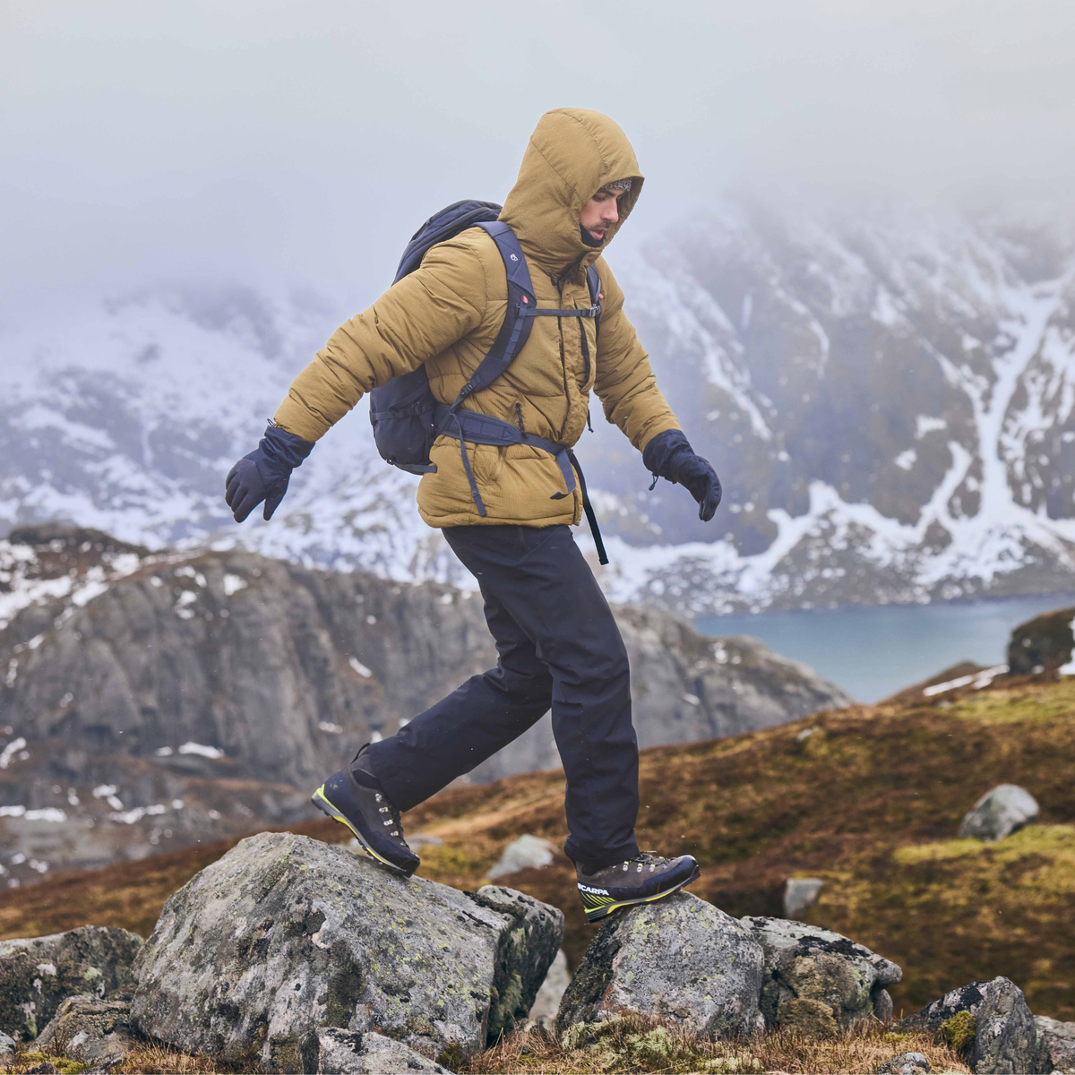 A man hiking up a rocky mountain slope wearing a tan hooded puffer jacket, Craghoppers Men's Steall Pro Thermo Waterproof Trousers Black 800 CMW847, gloves, and a backpack, with snowy mountains and a body of water in the background.