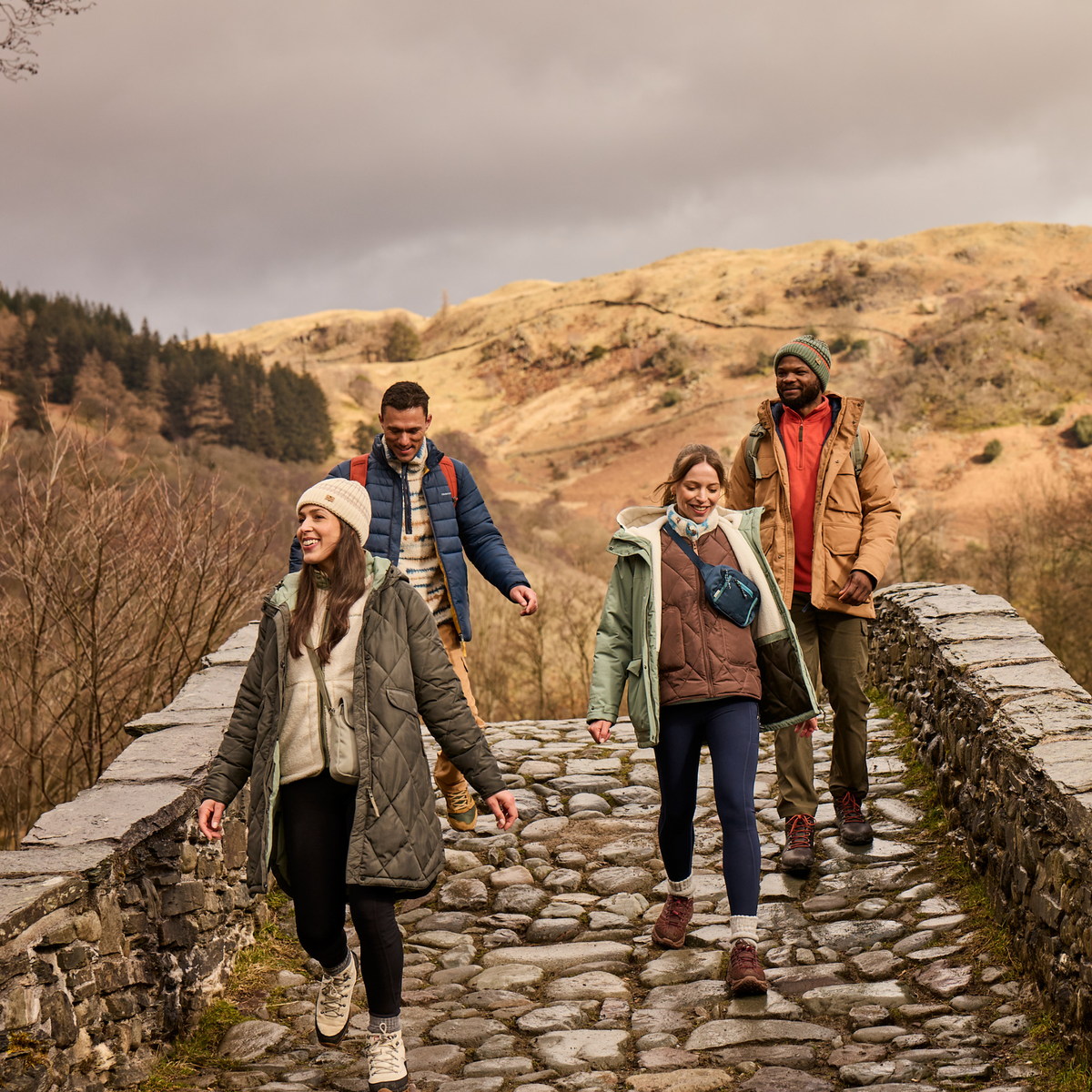 Four people walking along a stone path in a mountainous outdoor setting under an overcast sky, dressed in casual outdoor jackets and boots. One woman wearing Craghoppers Women’s Sweetgrass National Trust Insulated Parka Nori Green 1KZ CWN309