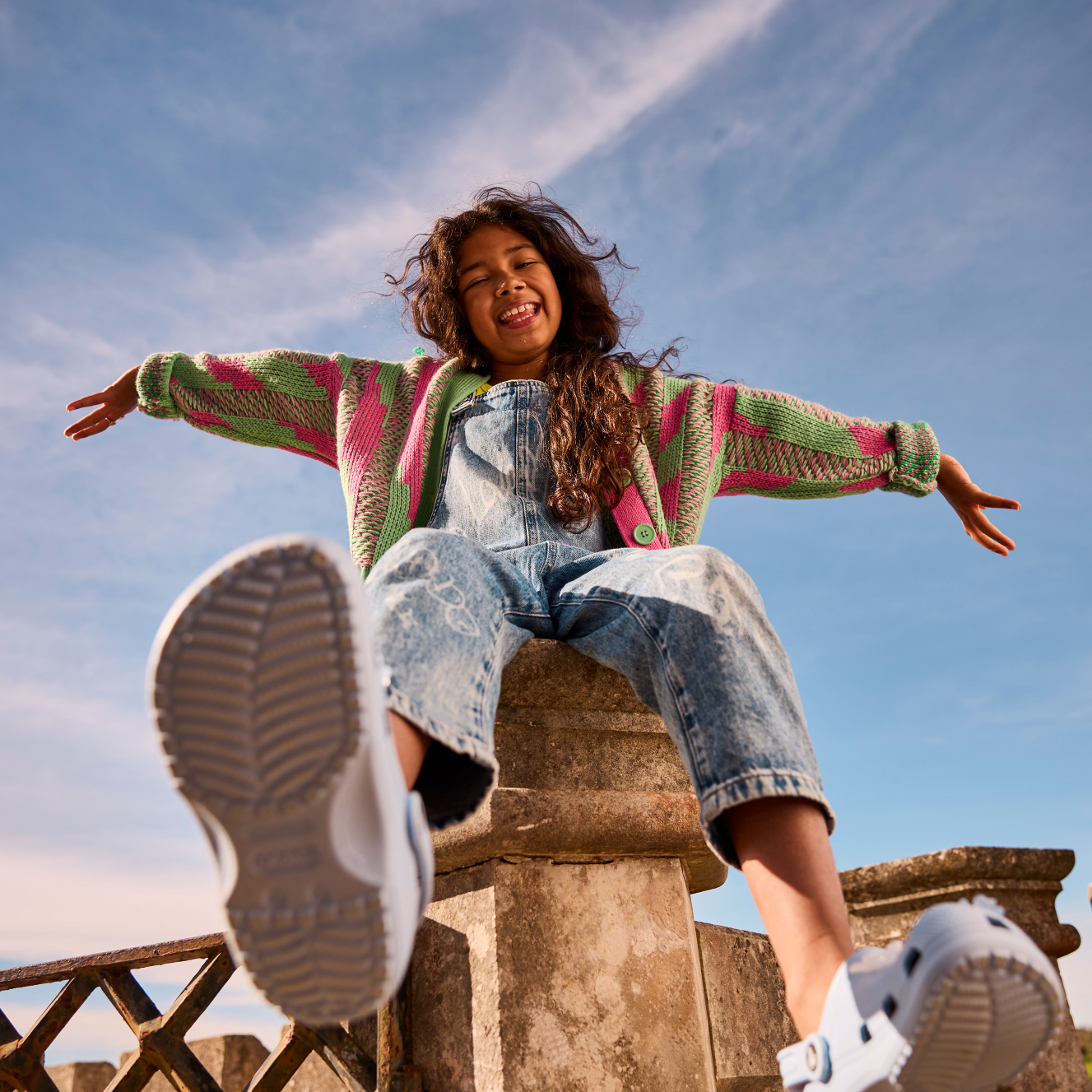 Young girl wearing Crocs Classic Clogs Junior Blue Calcite 4NS 206991 sitting on a fence